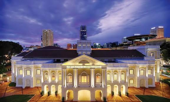 High-angle evening view of the illuminated Arts House building in Singapore, showcasing its white Palladian colonial facade set against a purple and blue twilight sky.