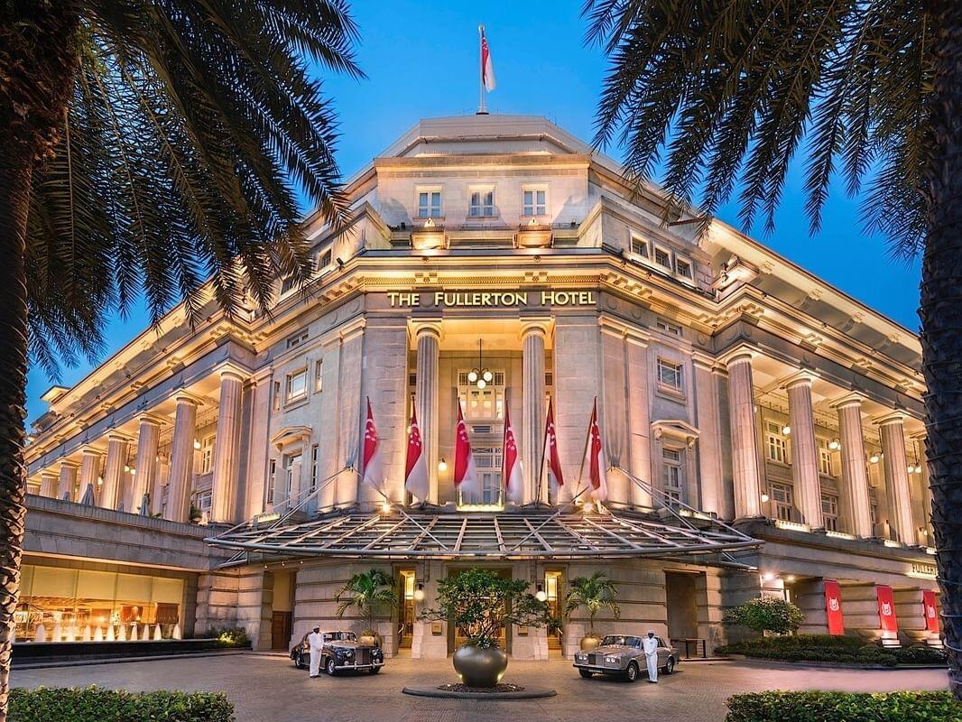 Low-angle evening view of the illuminated facade of The Fullerton Hotel Singapore, framed by palm trees with vintage cars parked at the grand entrance.