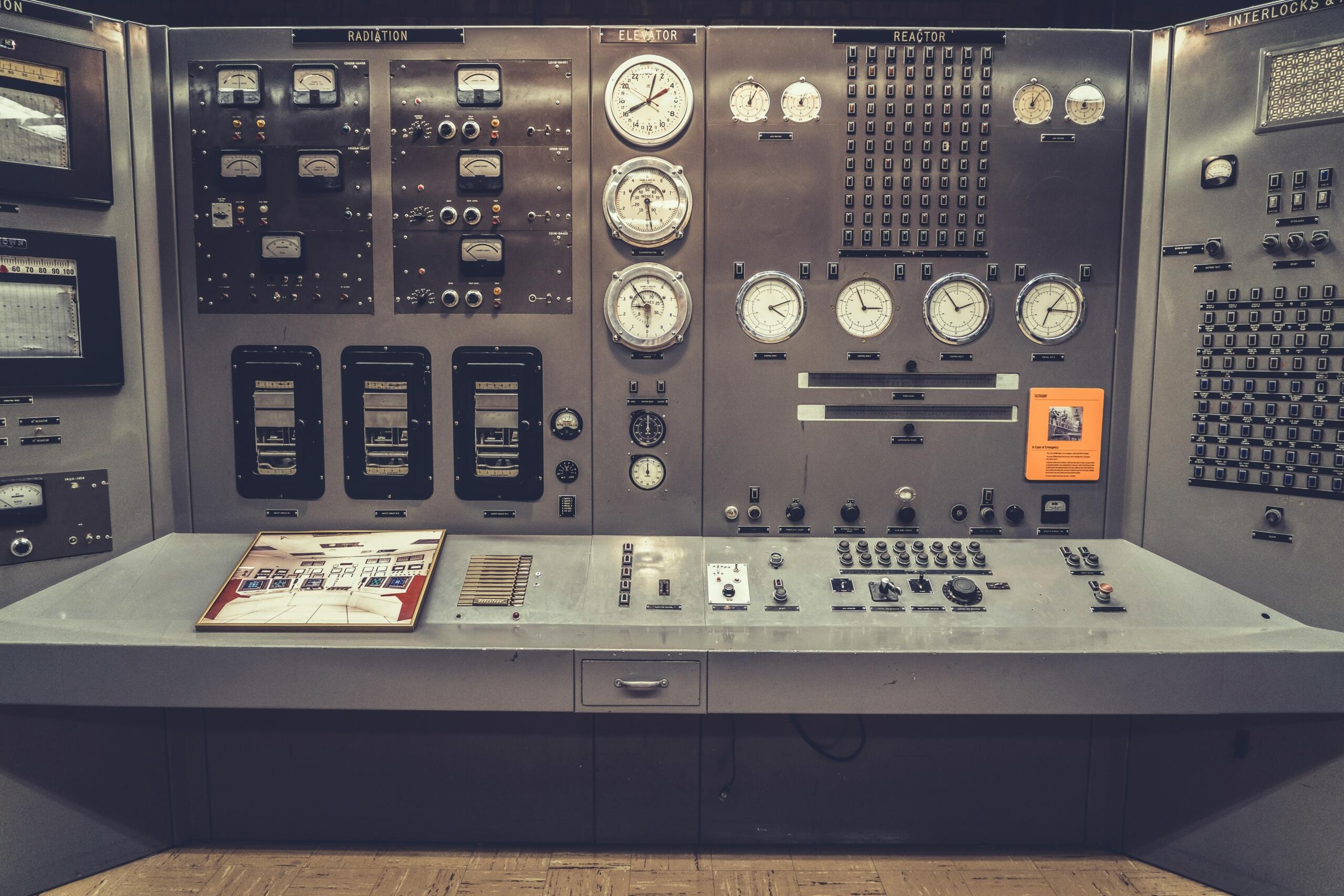 Slika prikazuje starinsko nadzorno ploščo, verjetno iz industrijskega ali znanstvenega okolja, kot je jedrska elektrarna ali laboratorij. Na plošči je veliko analognih merilnikov, stikal, gumbov in prikazovalnikov, ki služijo spremljanju in upravljanju različnih sistemov.