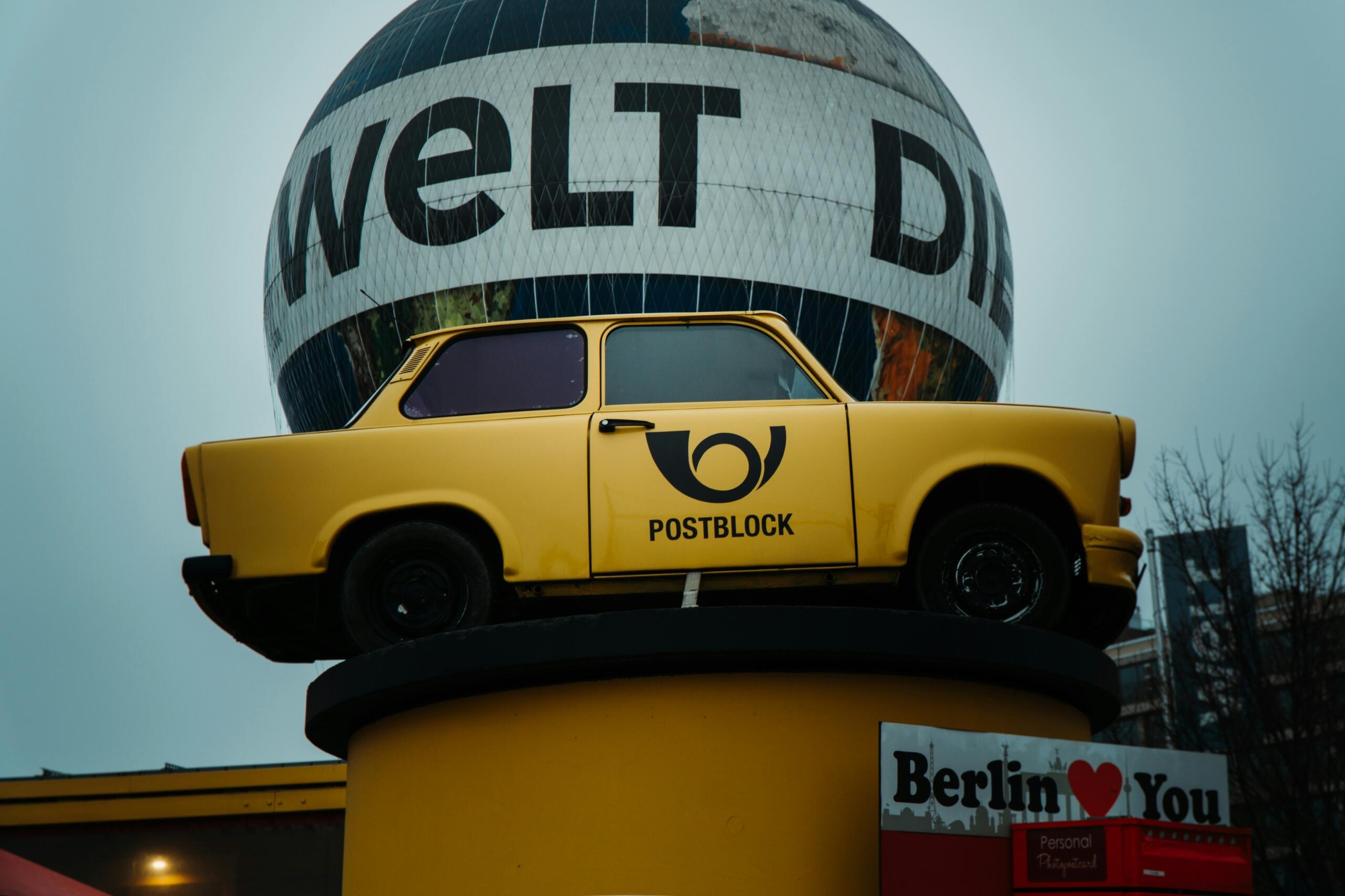 rumeni Trabant s poštnim logotipom in svetlobnim balonom Welt v ozadju v Berlinu