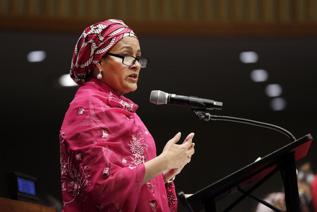 A person wearing a pink patterned top, a headscarf and glasses stands in front of a lectern bearing a microphone in an auditorium, addressing attendees. 