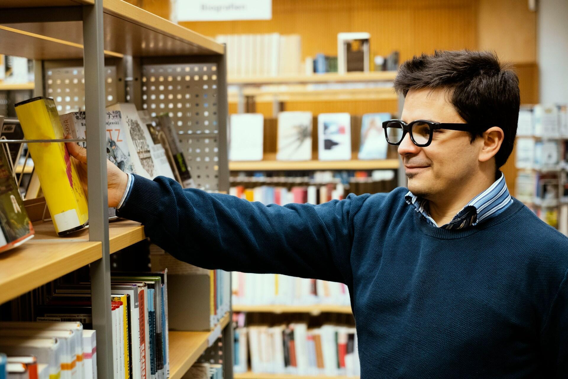 A man in a blue jumper takes a book from a shelf.