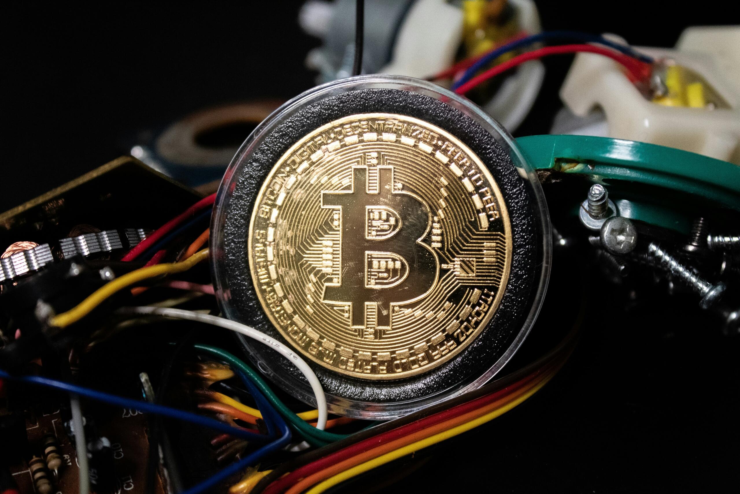 Zlati Bitcoin kovanec na ozadju barvnih žic, vijakov in vezij v temnem okolju.