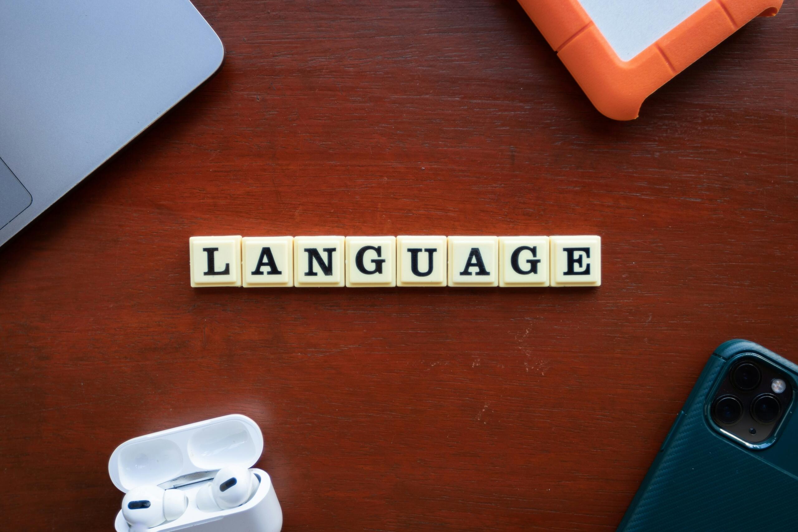 Lesene ploščice, ki tvorijo besedo 'LANGUAGE' na mizi z laptopom, tablico, telefonom in slušalkami.