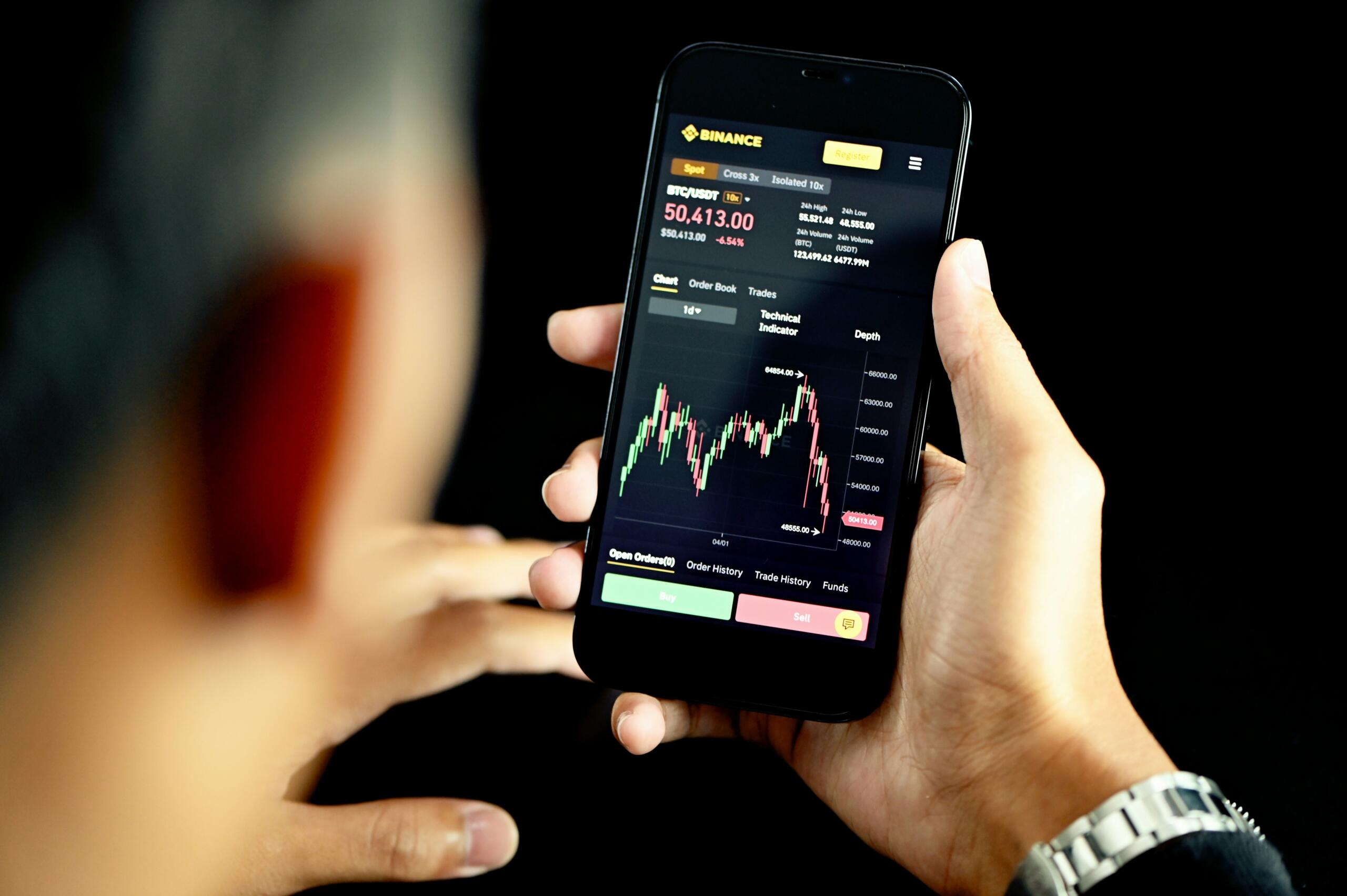 Oseba drži pametni telefon z aplikacijo Binance in grafikonom cene BTC/USDT v temnem okolju.