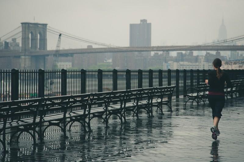 Ženska teče po mokri promenadi v dežju z Brooklyn Bridgeom v ozadju, New York Podpis fotografije: Redna telesna aktivnost, tudi v slabem vremenu, izboljša prekrvavitev možganov in pomaga pri boljšem učenju jezikov. Foto: Garry Knight