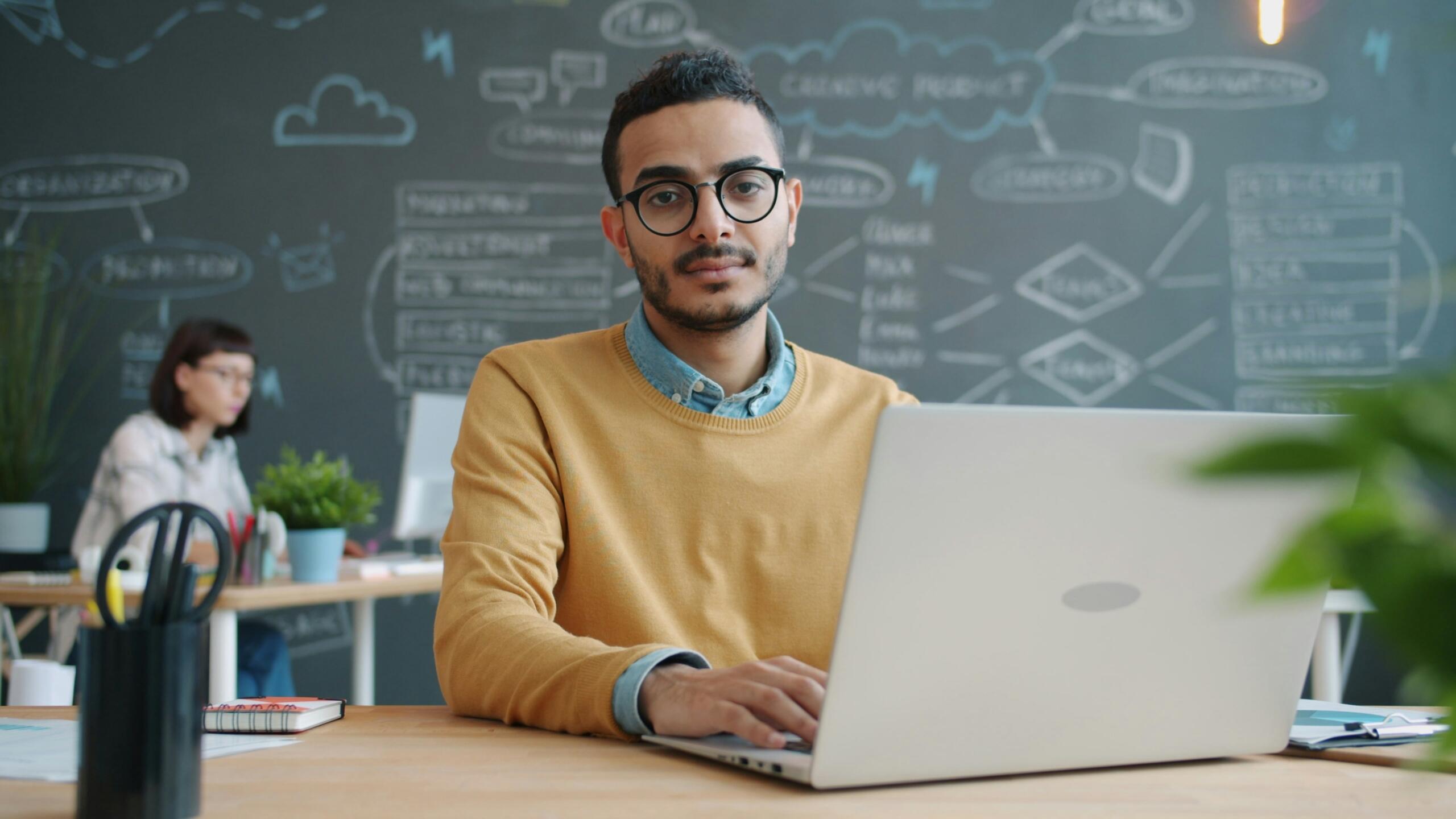 Mlajši moški v sodobni pisarni dela na prenosniku, za njim je tabla z diagrami in zapiski.