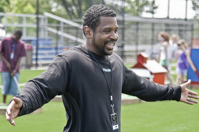 Nasmejan moški trener v črni majici z razširjenimi rokami na športnem igrišču na prostem