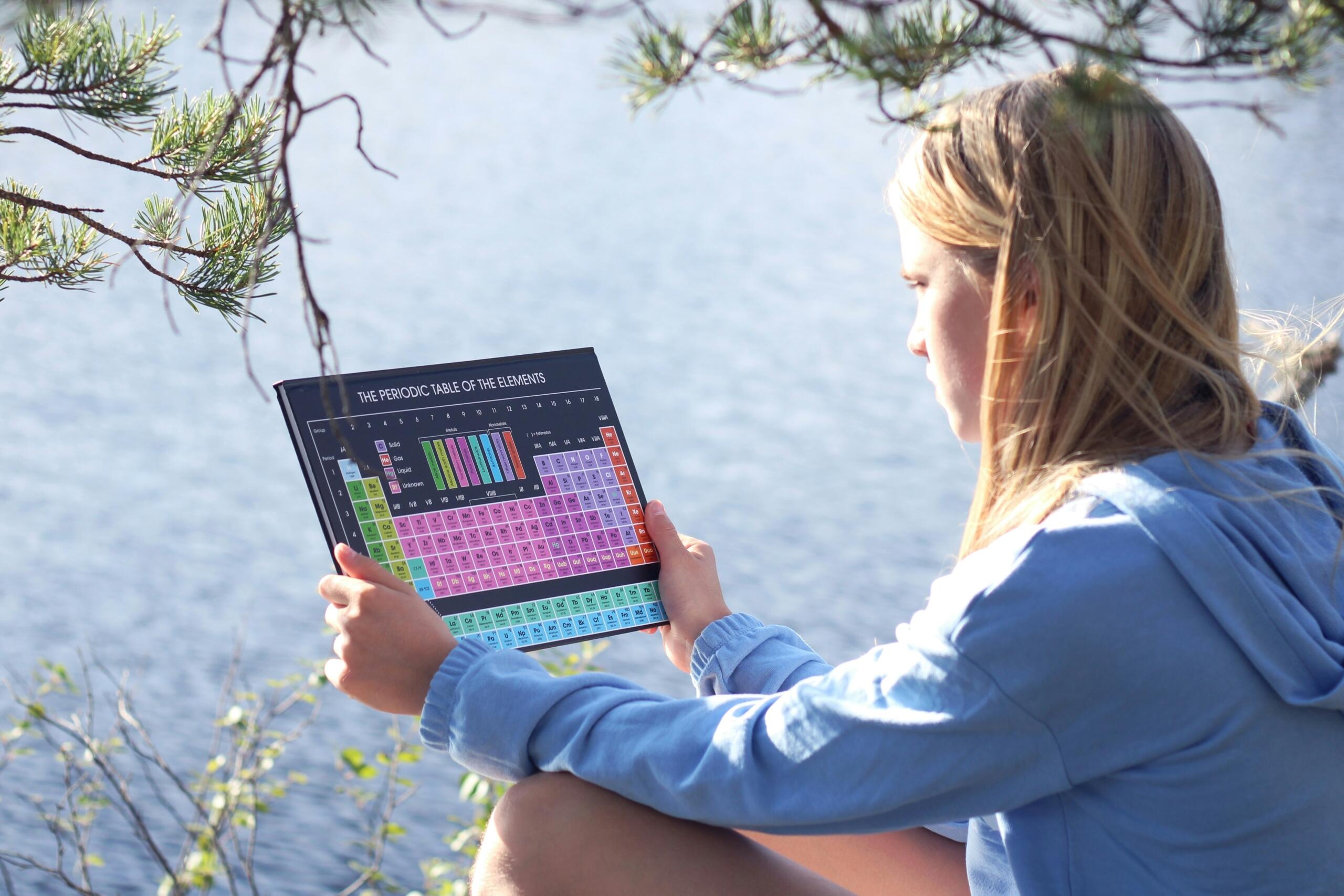 Mlada deklica v modri kapuci sedi na skali ob jezeru, drži velik barvni periodni sistem elementov v rokah, ozadje z borovci in vodo