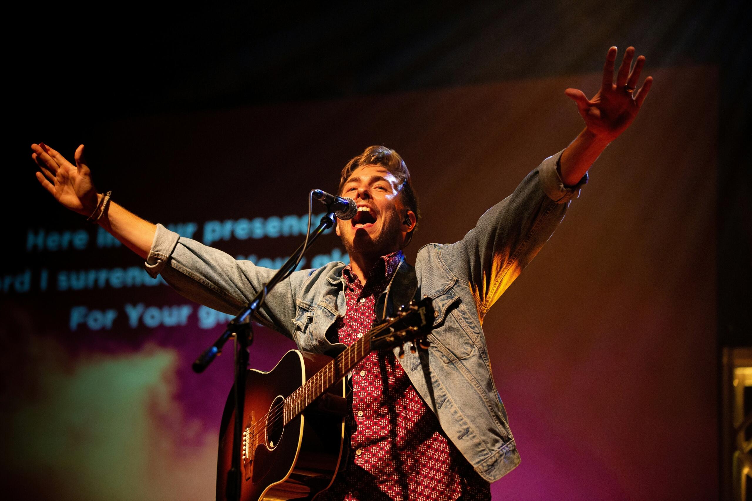 Moški z rjavimi lasmi v denim jakni strastno poje v mikrofon, drži akustično kitaro in razširja roke na odru z osvetljenim ozadjem