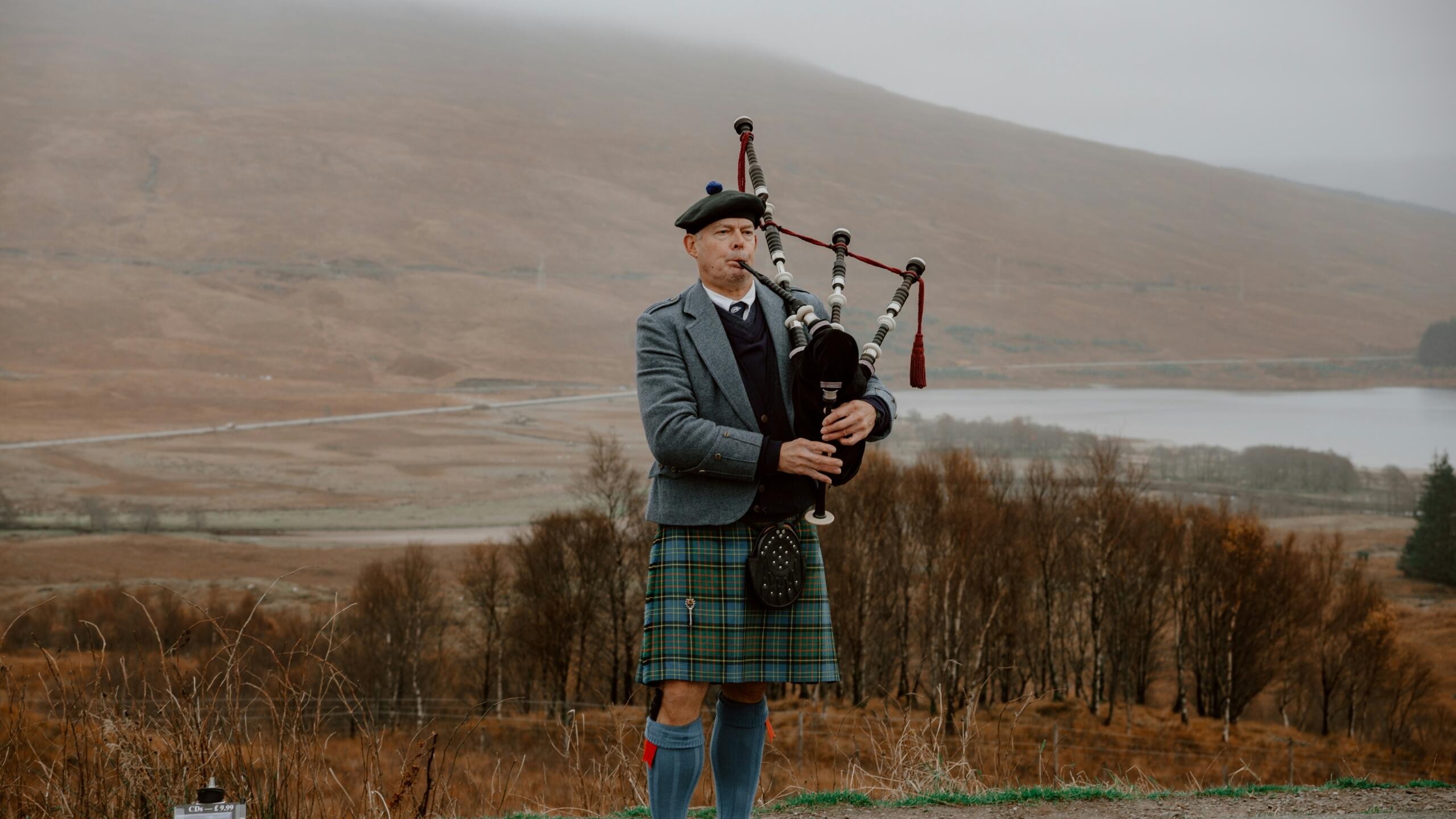 Moški v tradicionalni škotski obleki igra na gajde na hribu z jezerom in meglico v ozadju