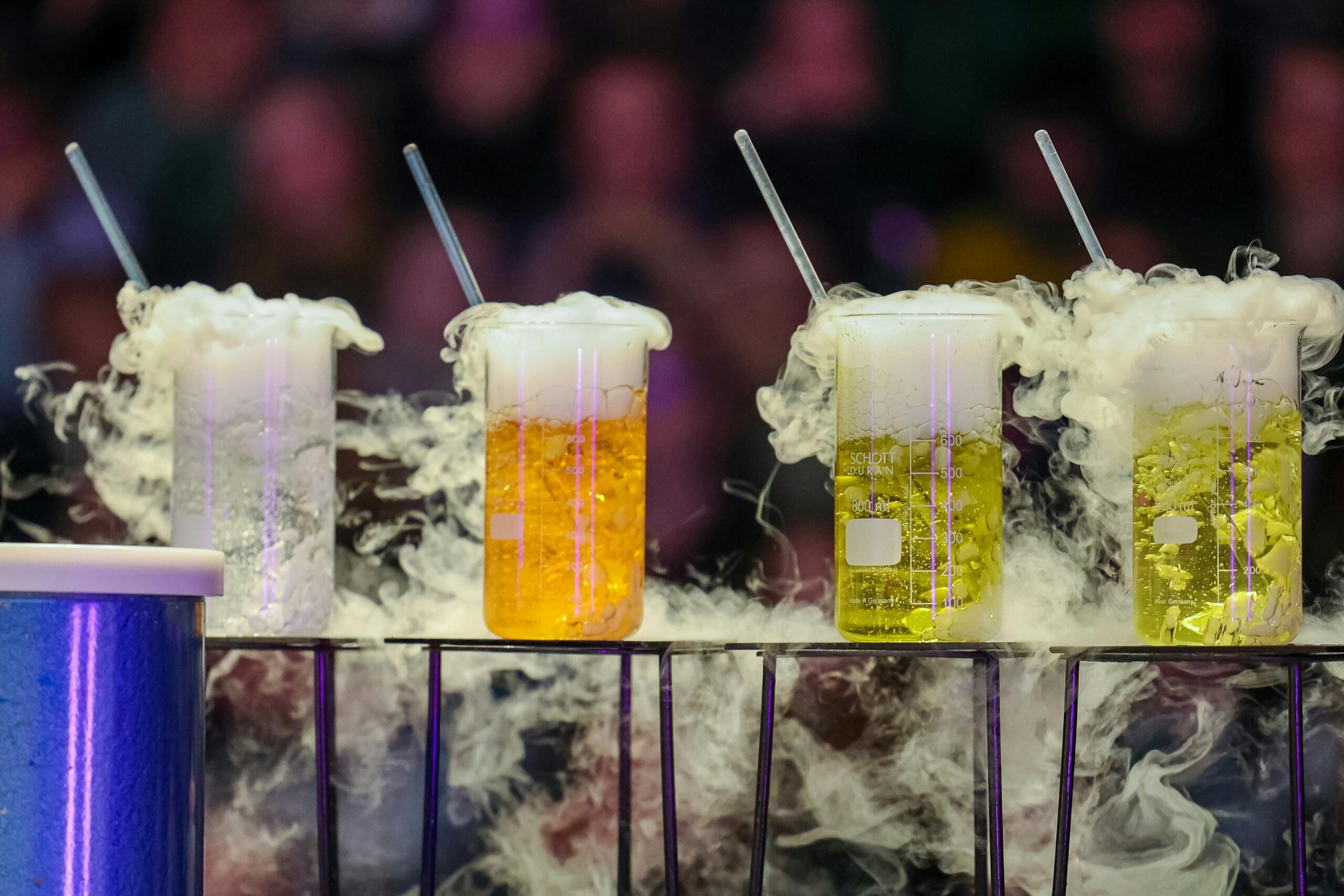 Štiri steklene posode na modri mreži v temnem laboratoriju, polne živahno oranžne in rumene tekočine z močnim belim dimom, ki se vali navzgor, steklene palčke potopljene v tekočino, ozadje zamegljeno z barvnimi lučmi in množico ljudi