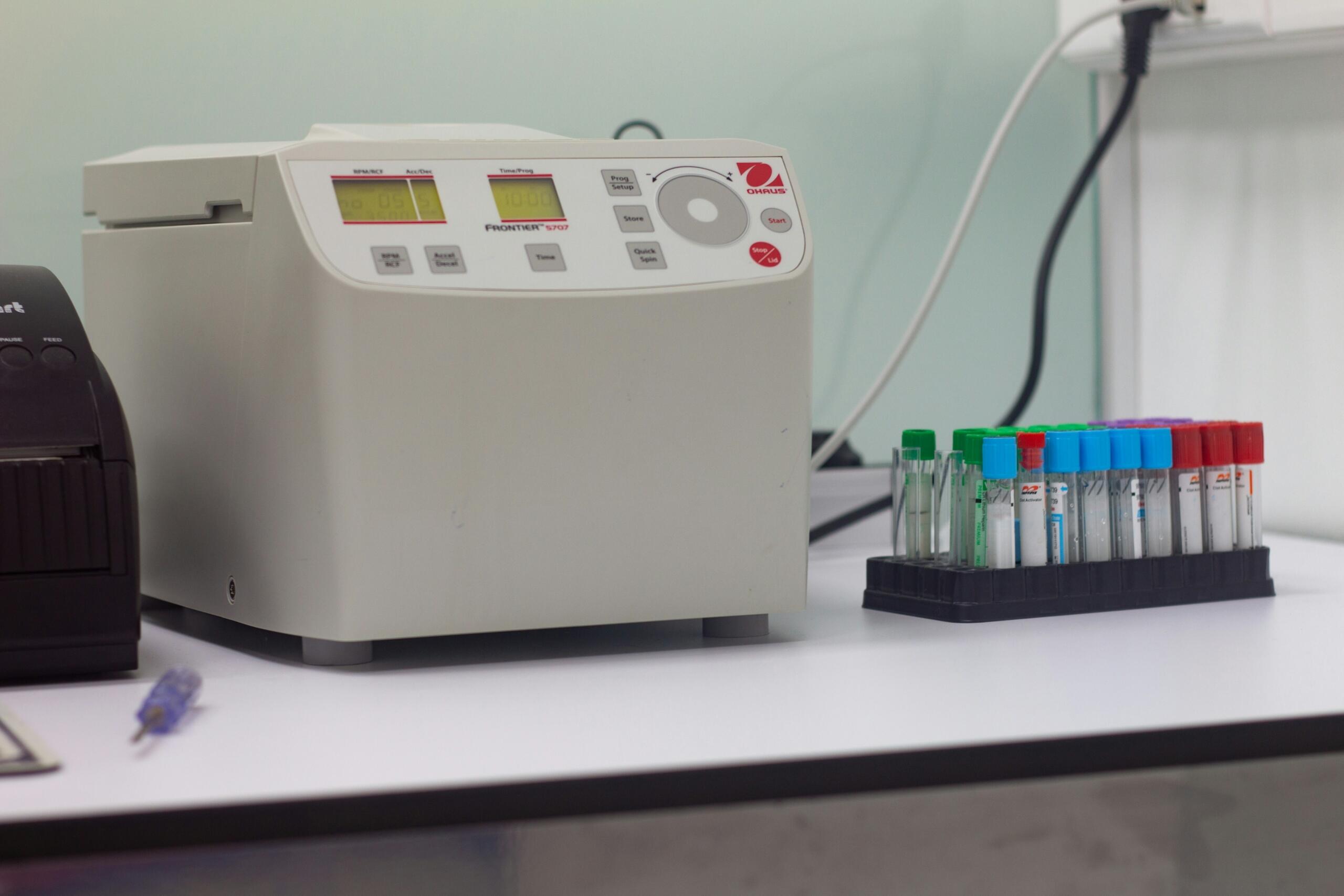 Bela centrifuga OHAUS Frontier 5707 na laboratorijski mizi z digitalnim zaslonom, gumbi za nastavitev hitrosti (RPM/RCF), časom in programa, ob strani stojalo z epruvetami v barvnih pokrovih (zeleni, rdeči, modri, vijolični), ozadje zamegljeno z drugo opremo