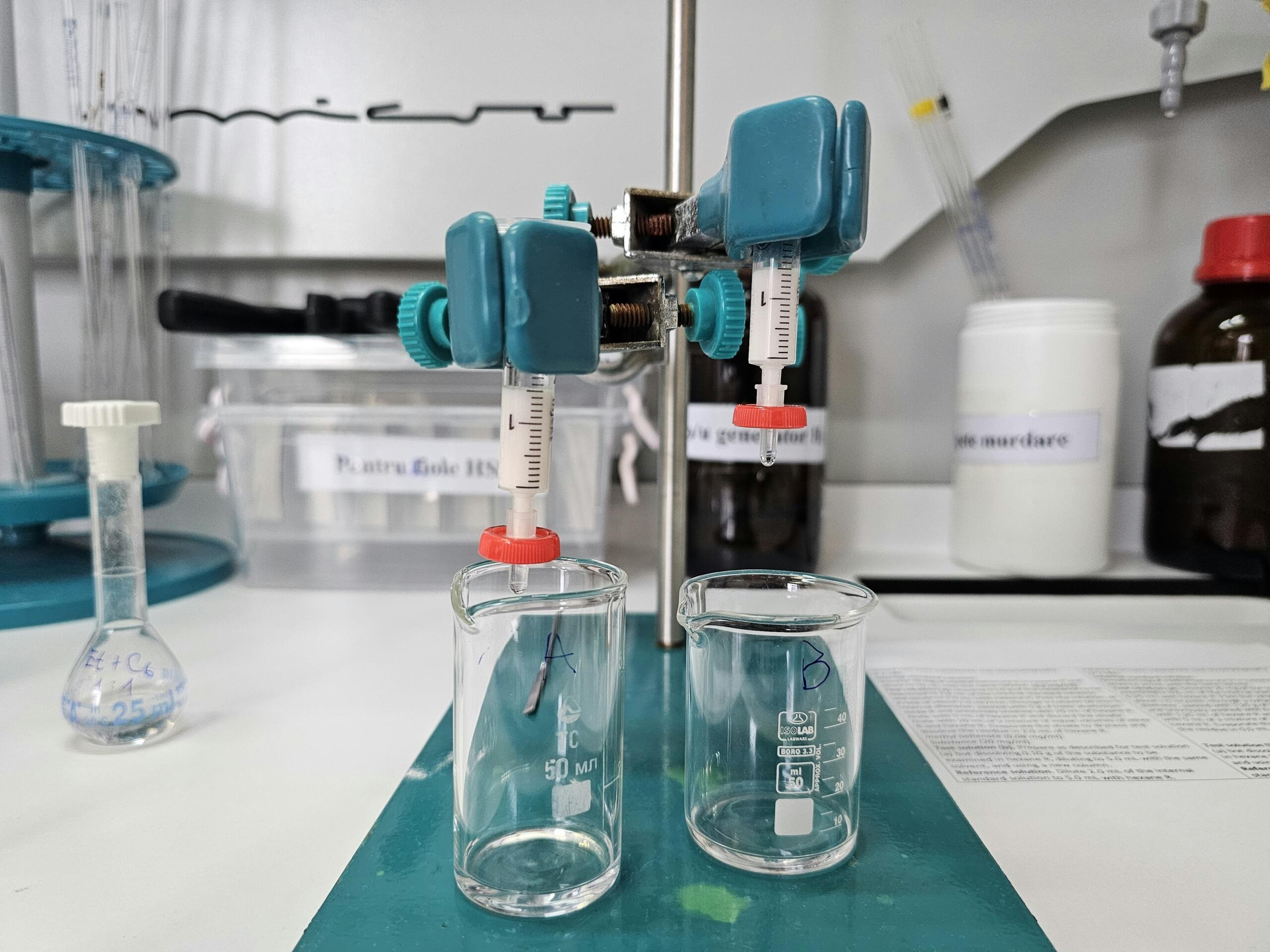 Stojalo drži elektronsko pipeto in kaplja majhno količino tekočine v prozorno mikrocentrifugno cevko, pritrjeno navpično na stojalo s turkiznimi objemkami, ozadje zamegljeno z drugimi epruvetami in steklenicami