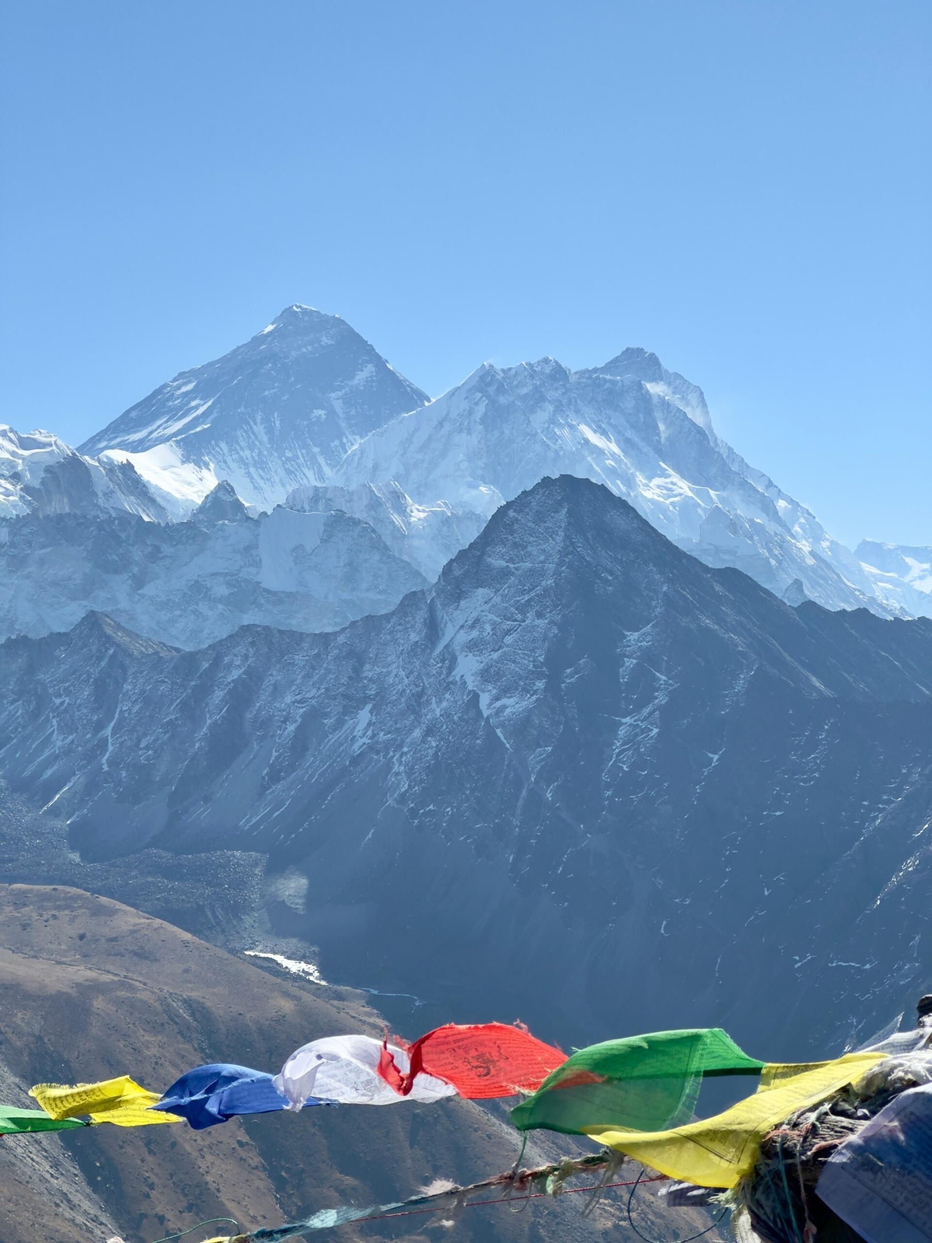Panoramski pogled na mogočni Mount Everest in okoliške himalajske vrhove pod jasnim modrim nebom, v ospredju barvne molitvene zastavice (rdeče, modre, zelene, rumene, bele) vihrajo na vetru na gorskem grebenu