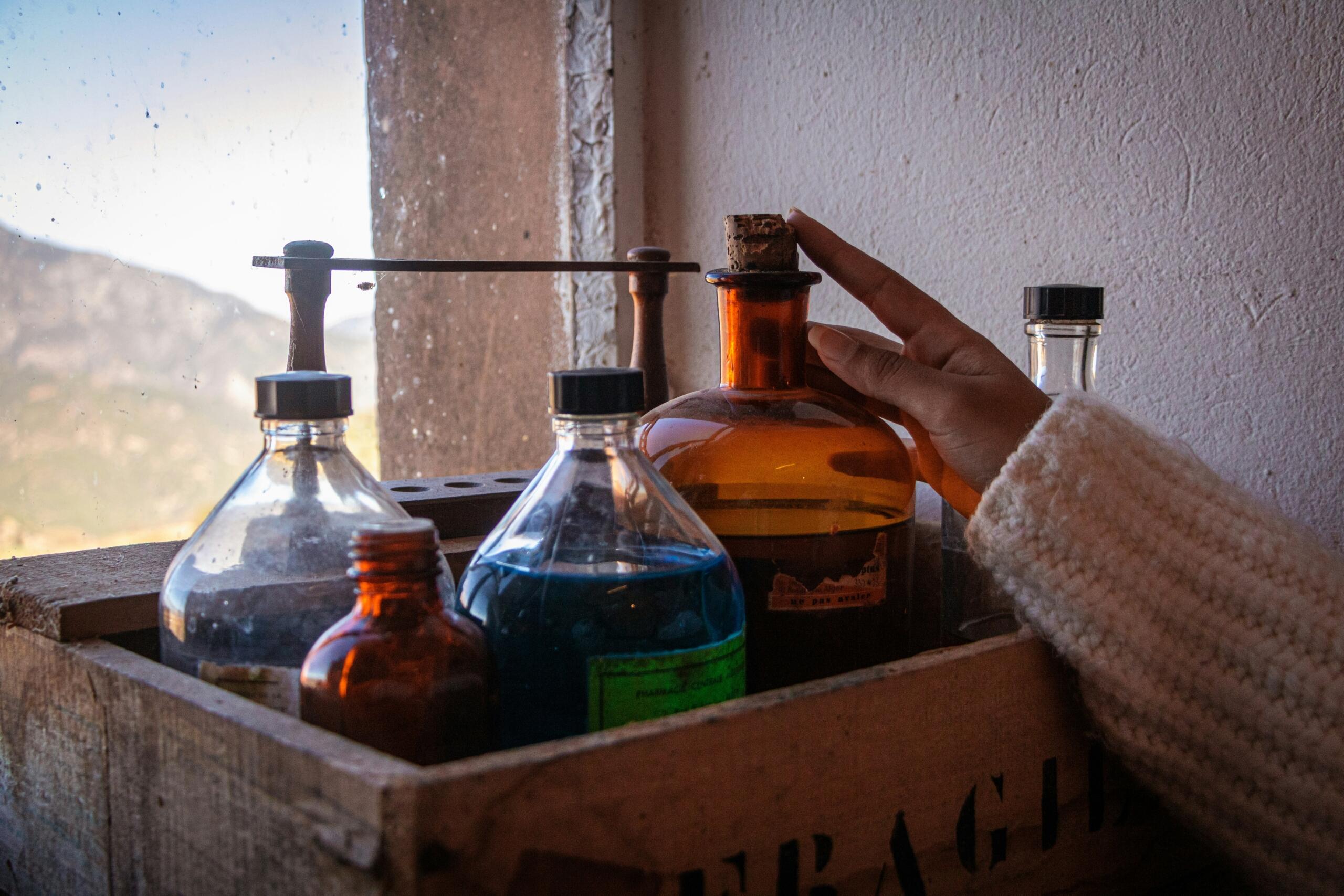 Ženska roka v svetlem puloverju drži staro janturno steklenico z modro tekočino iz lesenega zaboja z drugimi steklenicami, ozadje okno z razgledom na gore in nebo
