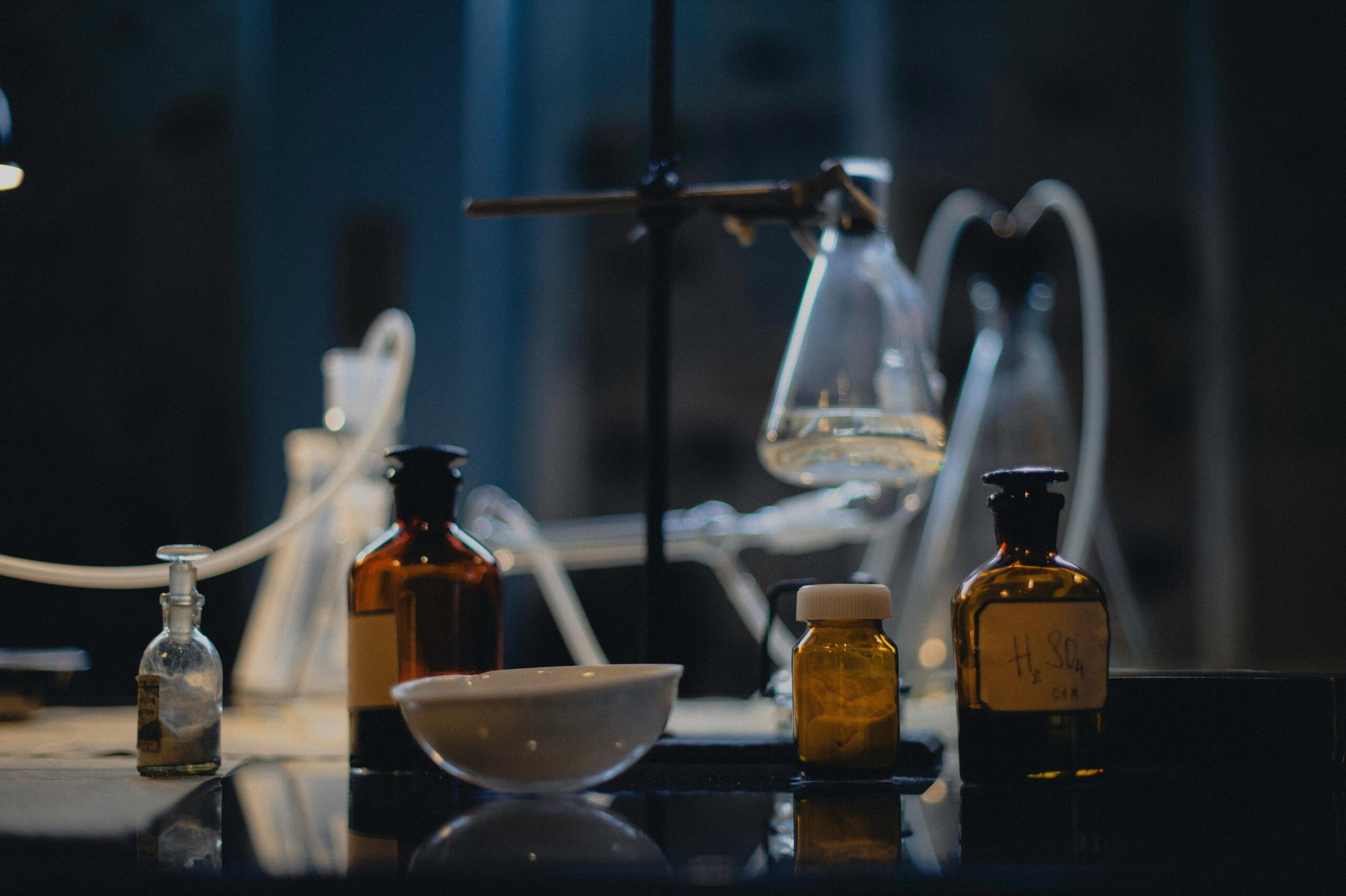 Temna laboratorijska miza z vintage steklenimi posodami, epruvetami in steklenicami z etiketami (npr. H₂SO₄), destilacijski aparat z gumijastimi cevmi, kristalna posoda in bakrena stativa v modri in oranžni osvetlitvi