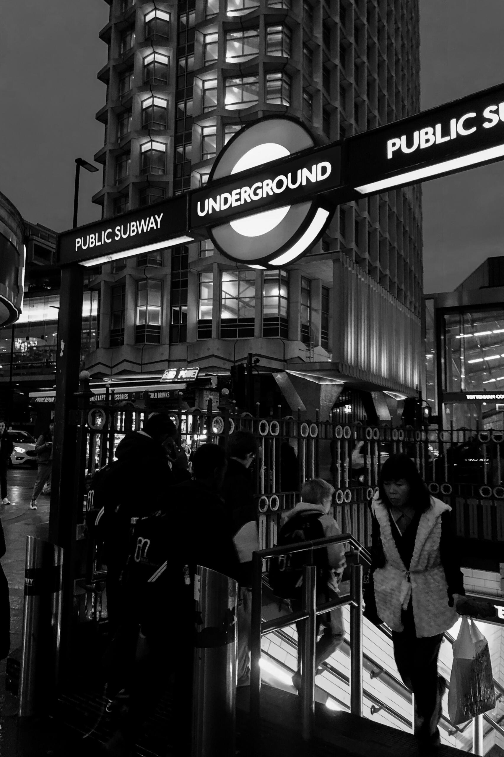 Črno-beli posnetek vhoda v londonski metro (Underground) z osvetljenim rdečim krogom in napisom "UNDERGROUND", nad njim moderna visoka stavba z osvetljenimi okni, mimoidoči na stopnicah in ograji