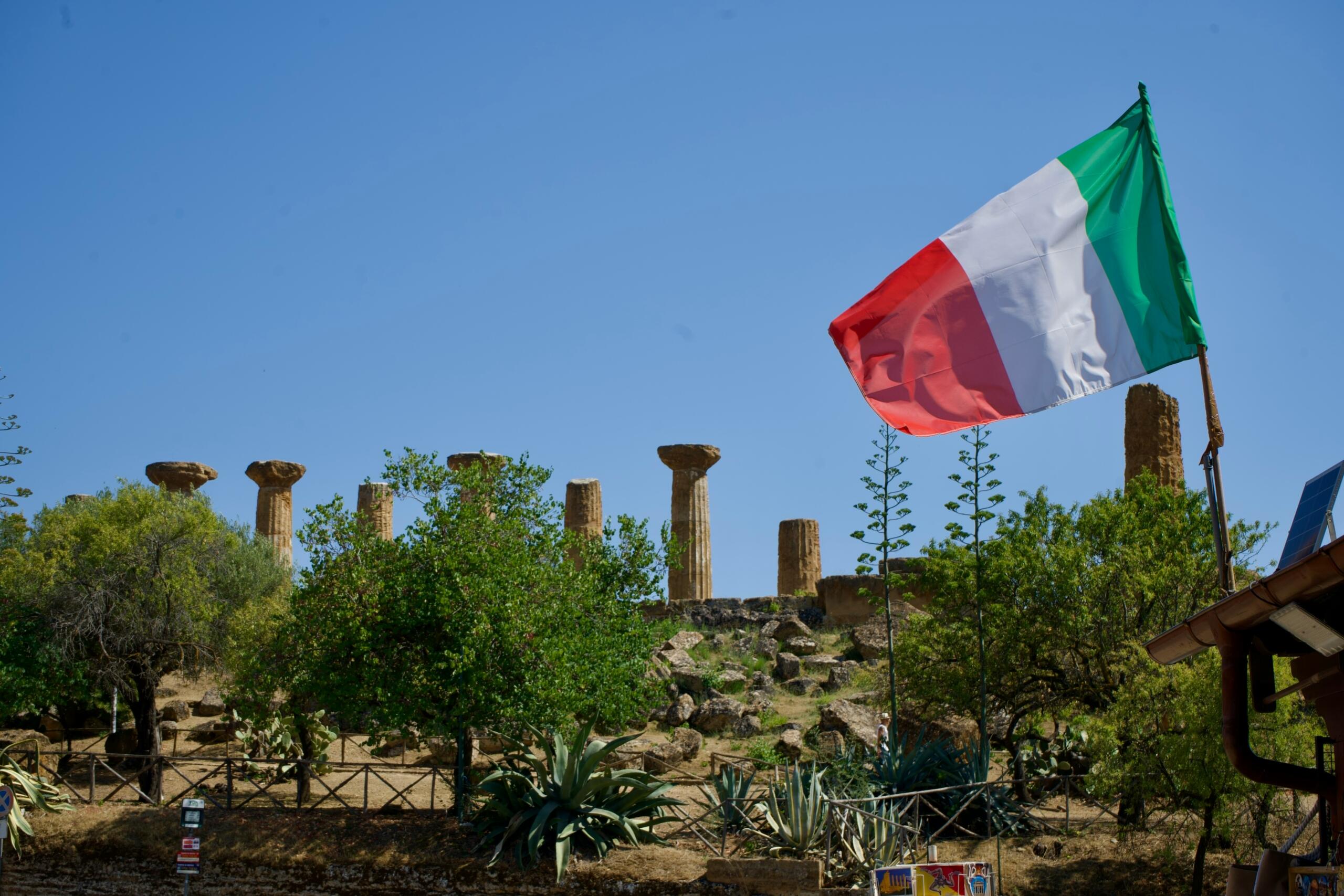 Italijanska zastava plapola pred antičnimi ruševinami v Italiji pod modrim nebom.