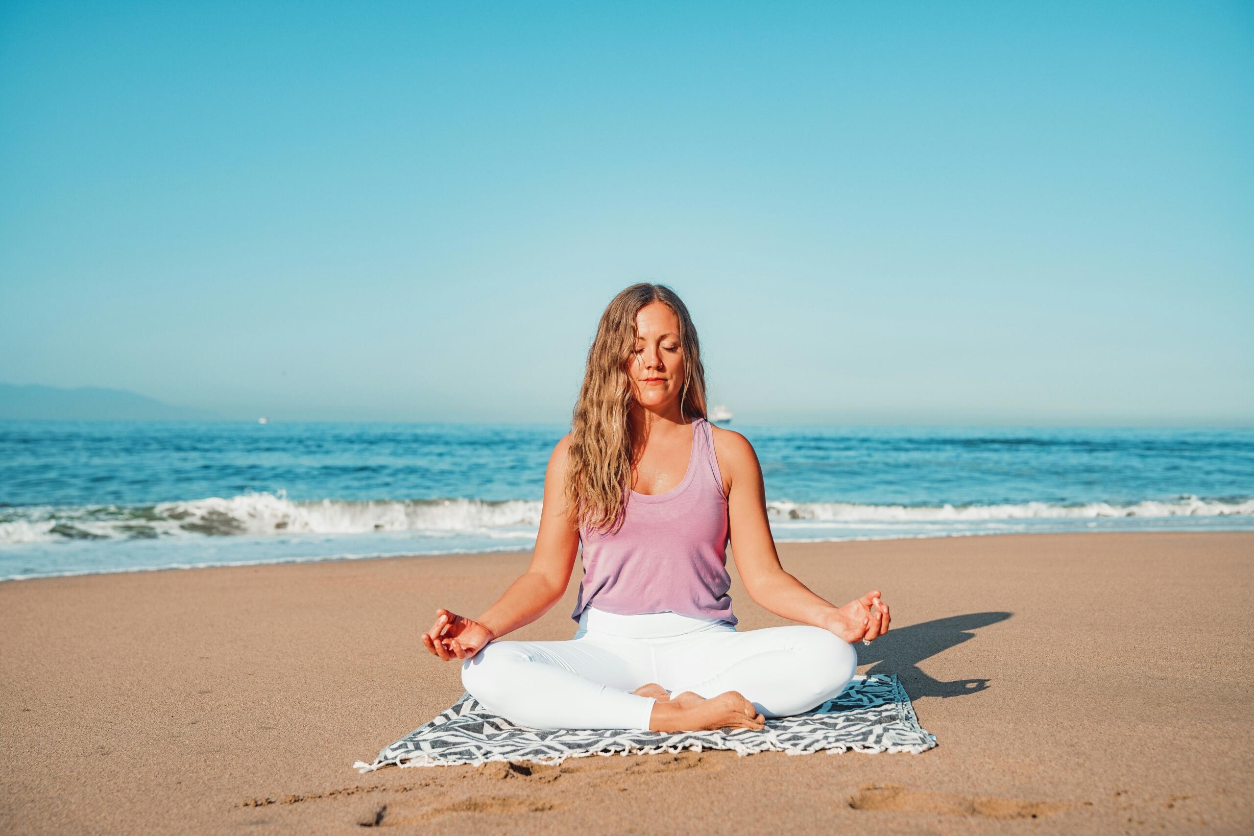 Ženska meditira na peščeni plaži ob morju v mirnem jutranjem vzdušju.