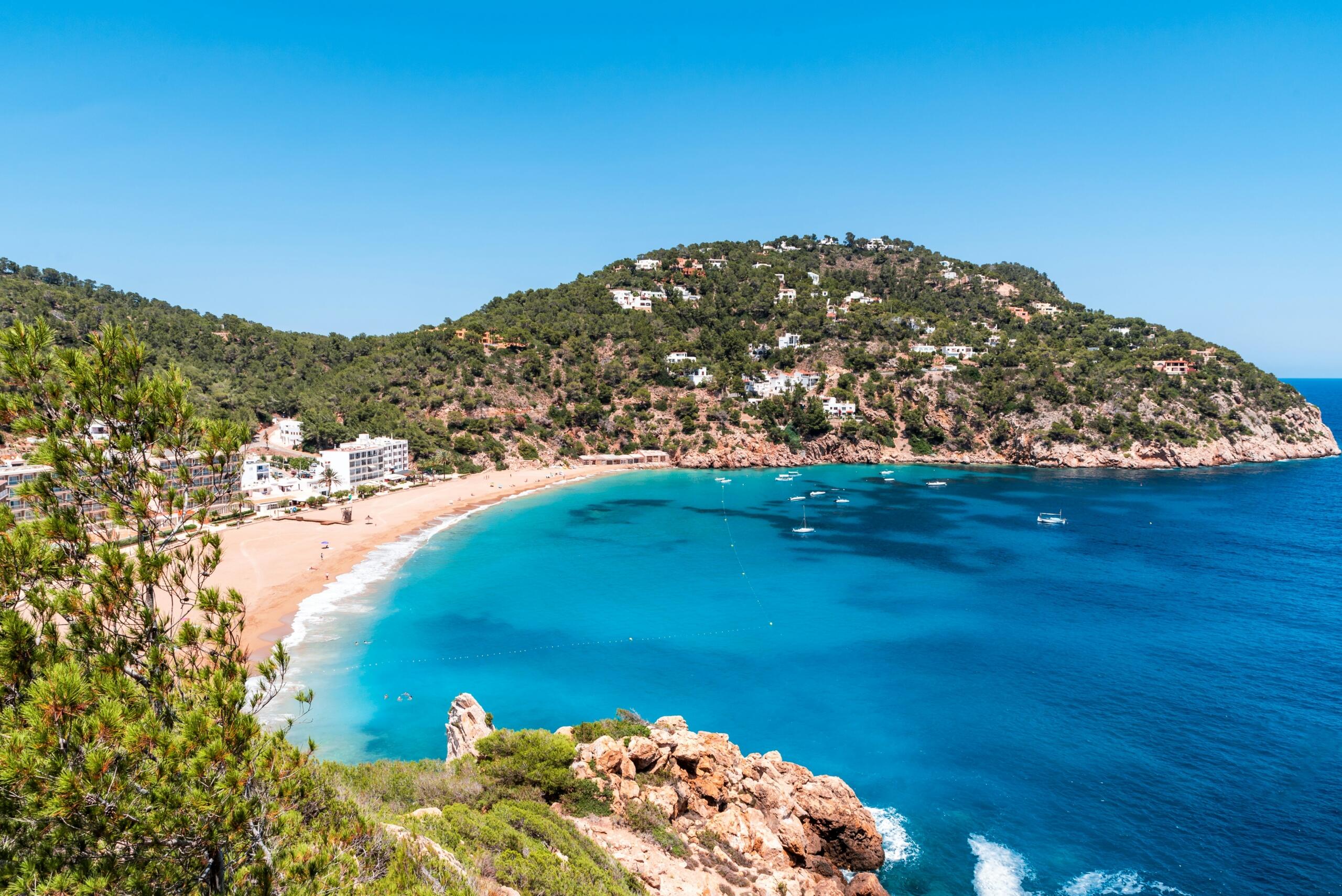 Panoramski pogled na zaliv Cala de Sant Vicent na Ibizi s turkizno modrim morjem, peščeno plažo in poraslim hribom z vilami