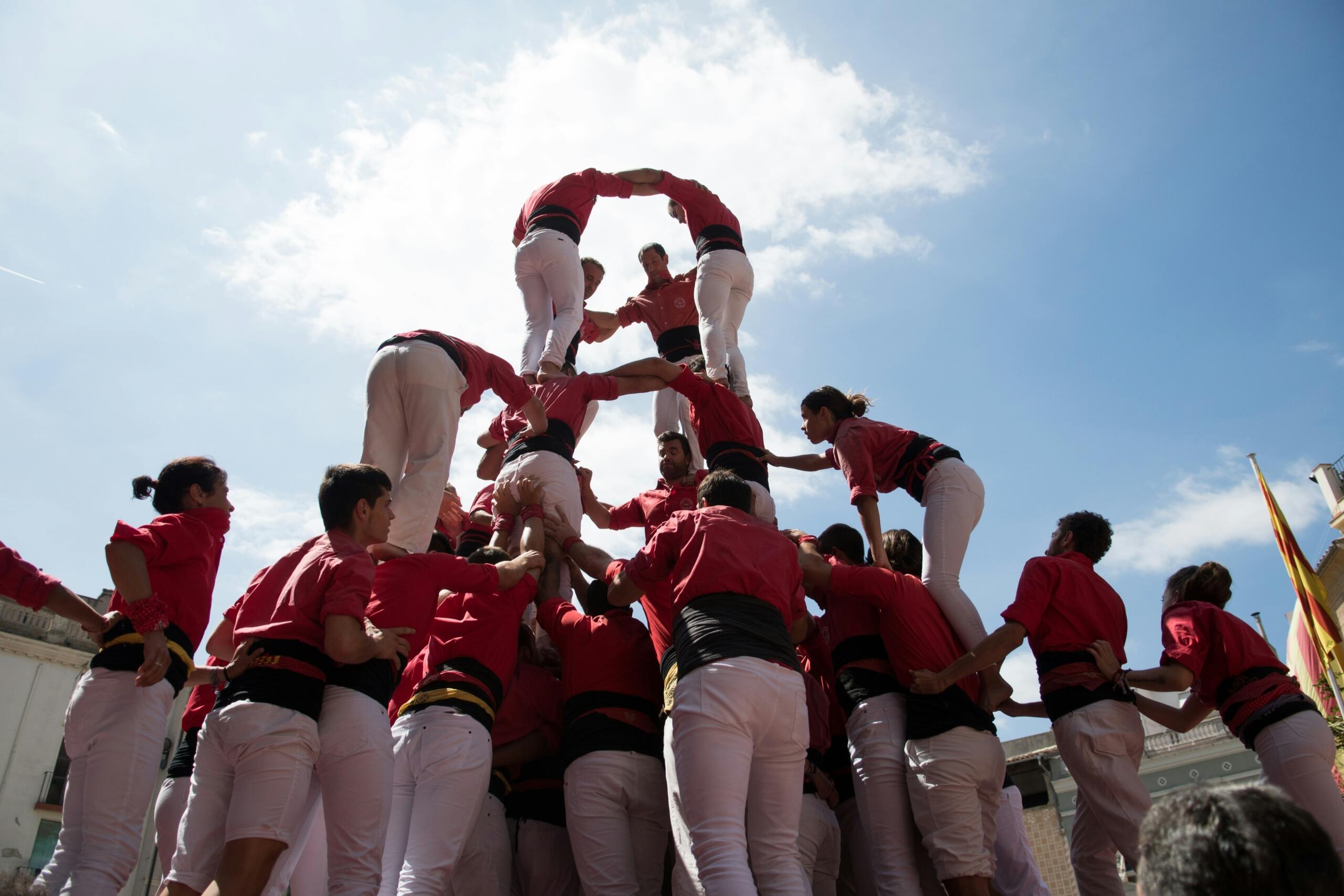 Skupina castellers v rdečih majicah in belih hlačah gradi človeški stolp na javnem trgu pod modrim nebom v Kataloniji