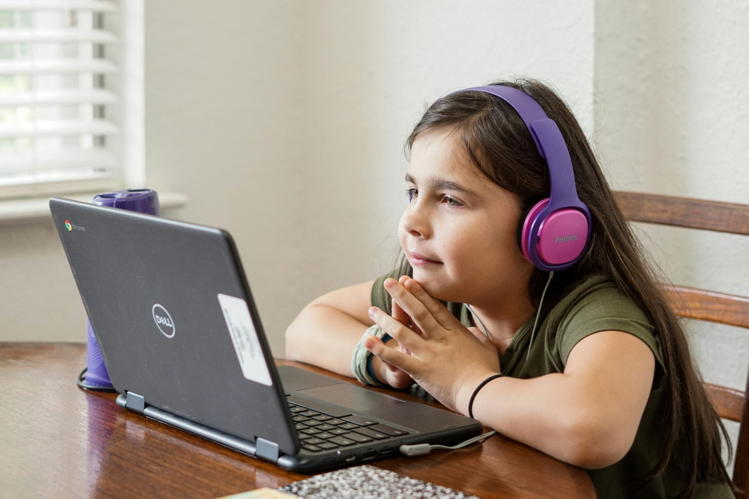 Mlajša deklica s temnimi dolgimi lasmi in vijoličnimi slušalkami Philips sedi za mizo pred Dell Chromebook prenosnikom in zbrano posluša spletni pouk