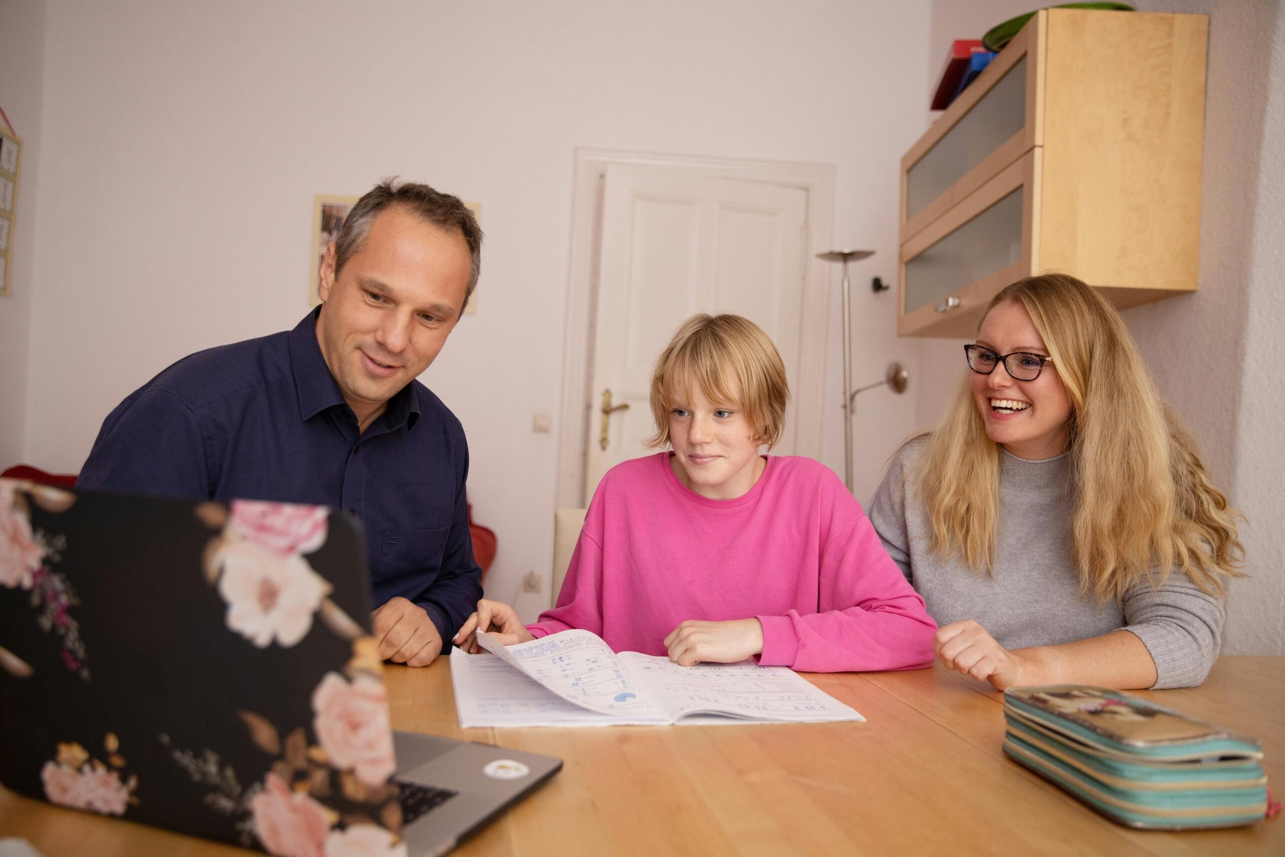 Oče v modri srajci in mama z blond lasmi in očali sedita ob sinu s svetlimi lasmi v rožnatem puloverju in skupaj pregledujeta zvezek pri leseni mizi