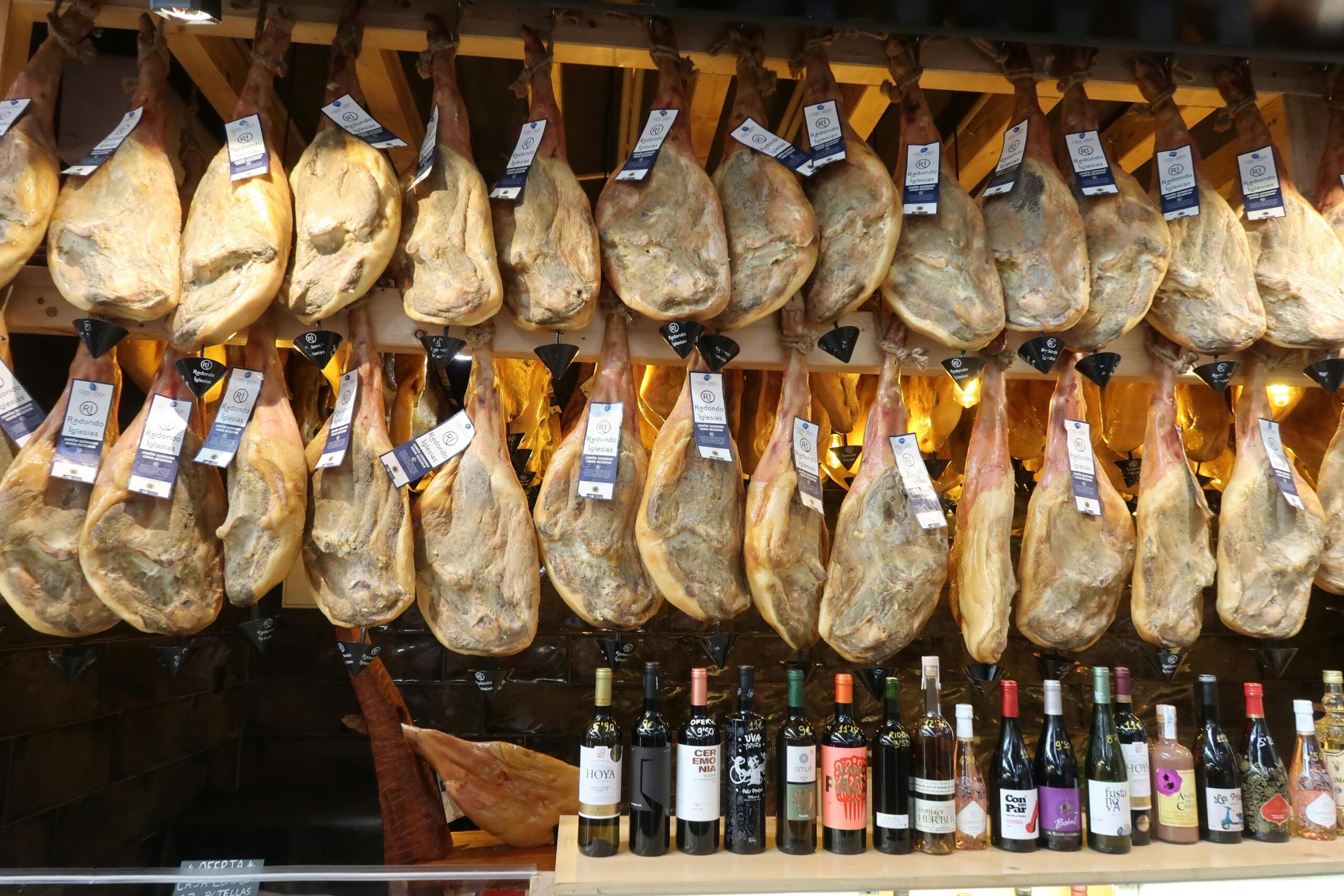 Številne noge sušenega šunke jamón ibérico visijo na lesenem stojalu v španski mesnici, spodaj vrsta steklenic španskega vina