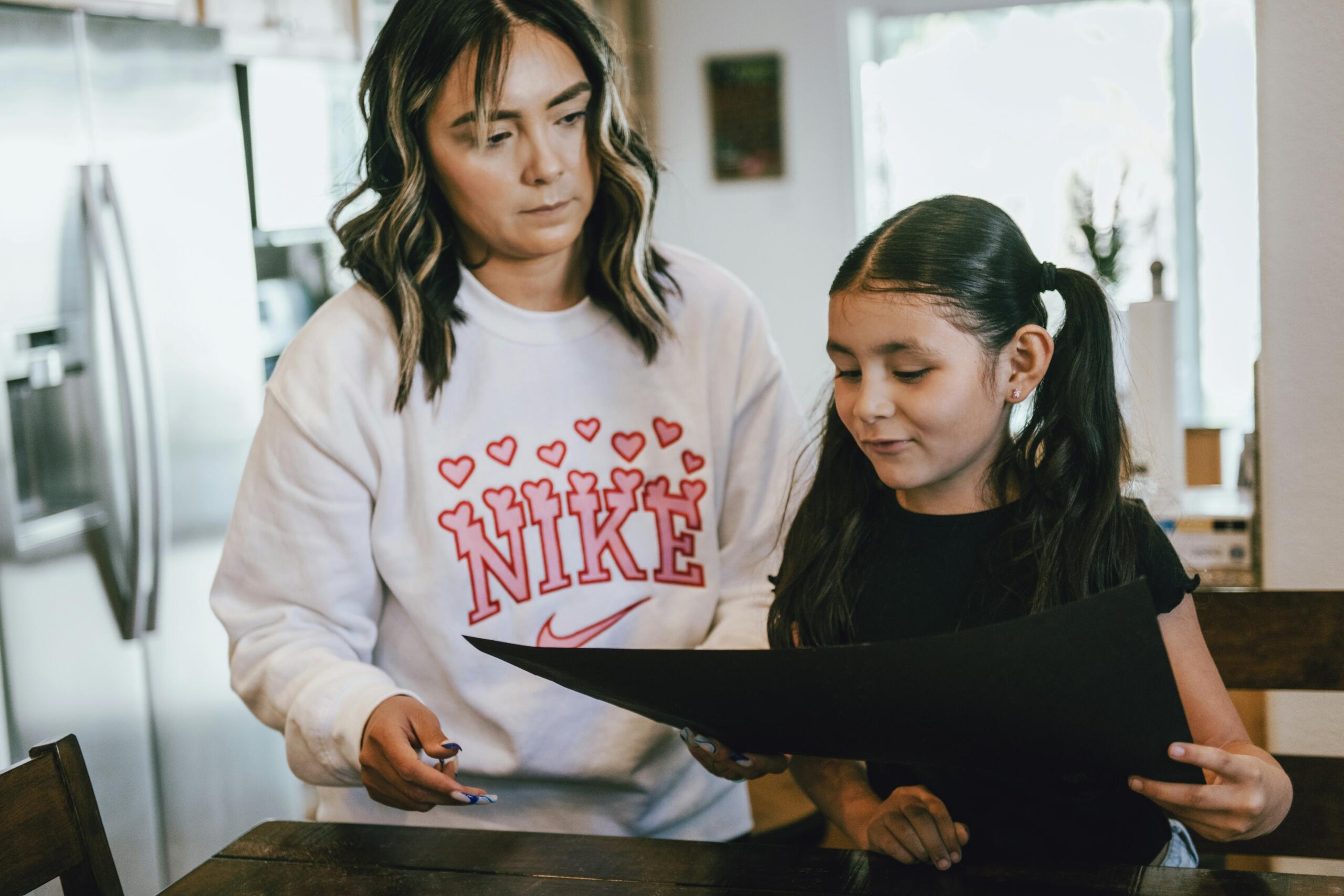 Mlada mama v belem Nike puloverju stoji za hčerko in skupaj gledata v odprto temno mapo z dokumenti pri kuhinjski mizi
