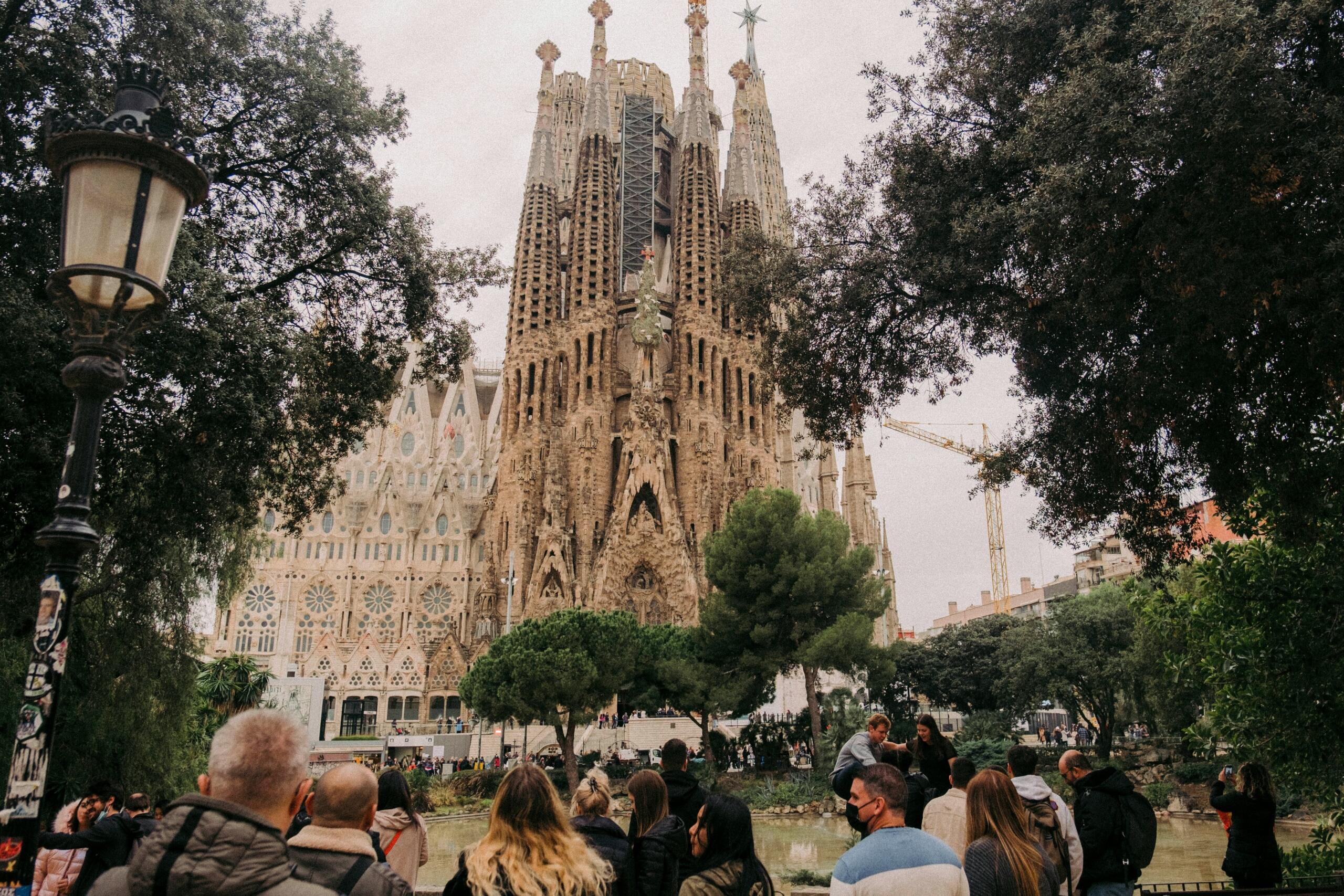 Množica turistov gleda Sagrado Famílijo Antonia Gaudíja iz parka z drevesi in ribnikom v ospredju