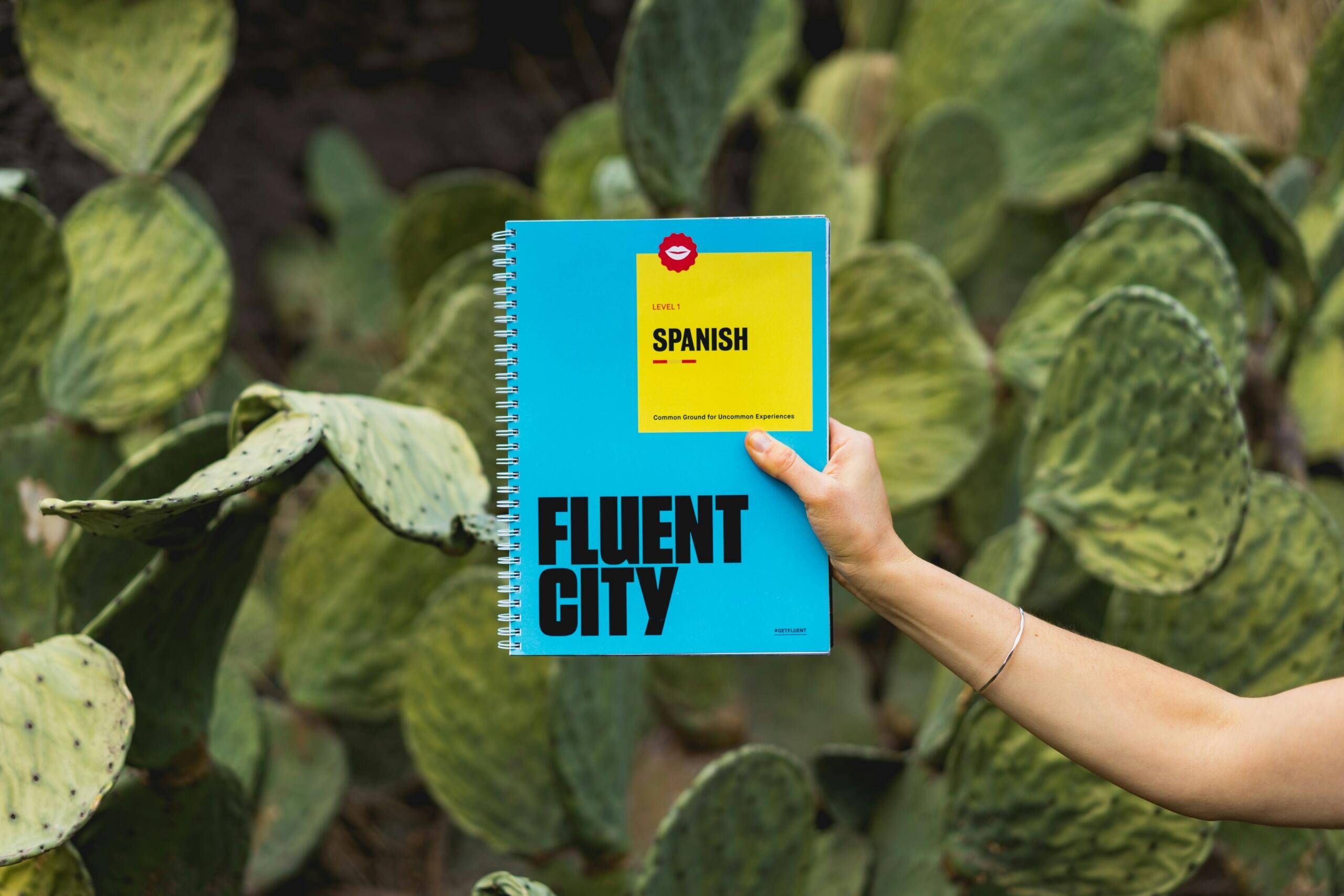 Ženska roka drži modro-rumeni spiralni učbenik španščine Level 1 znamke Fluent City pred zelenim kaktusovim ozadjem
