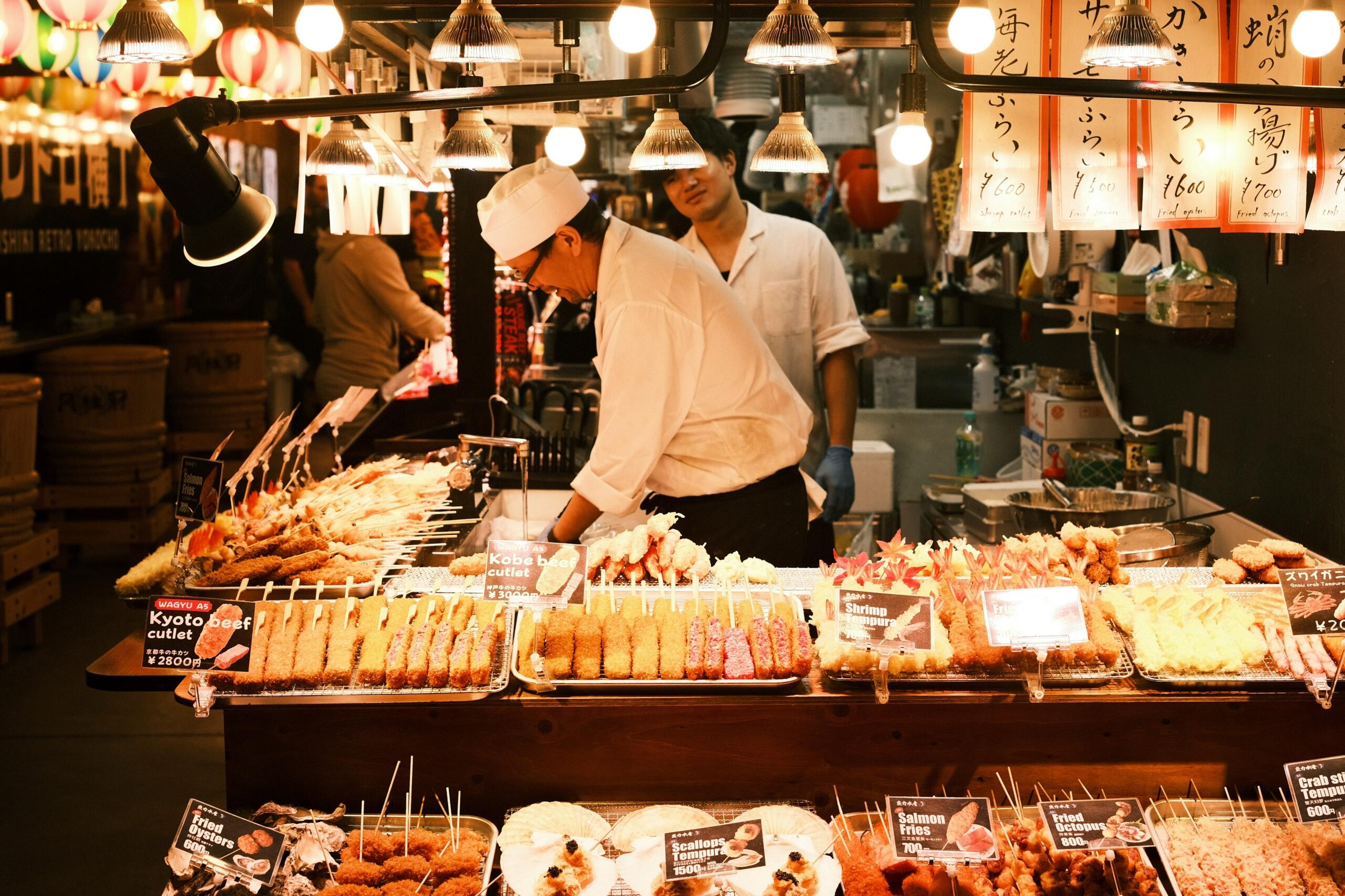Živahen japonski ulični stal z razstavljenimi ocvrtimi jedmi – katsu, tempura, ražnjiči, kuhar v beli uniformi pripravlja hrano pod svetlimi lučmi