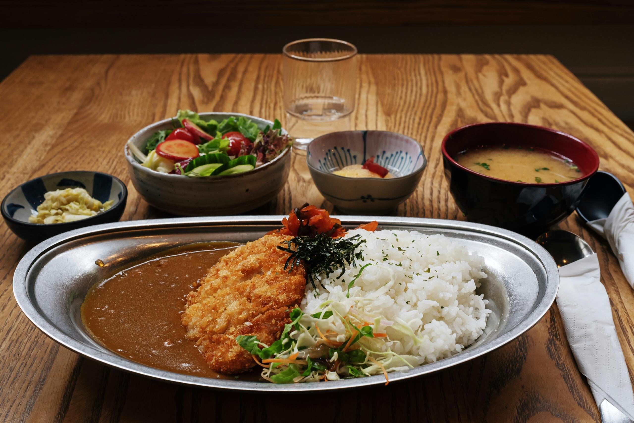 Kovinski pladenj s katsu curry – tonkatsu, bel riž, solata in temna omaka, ob njem miso juha in sveža solata
