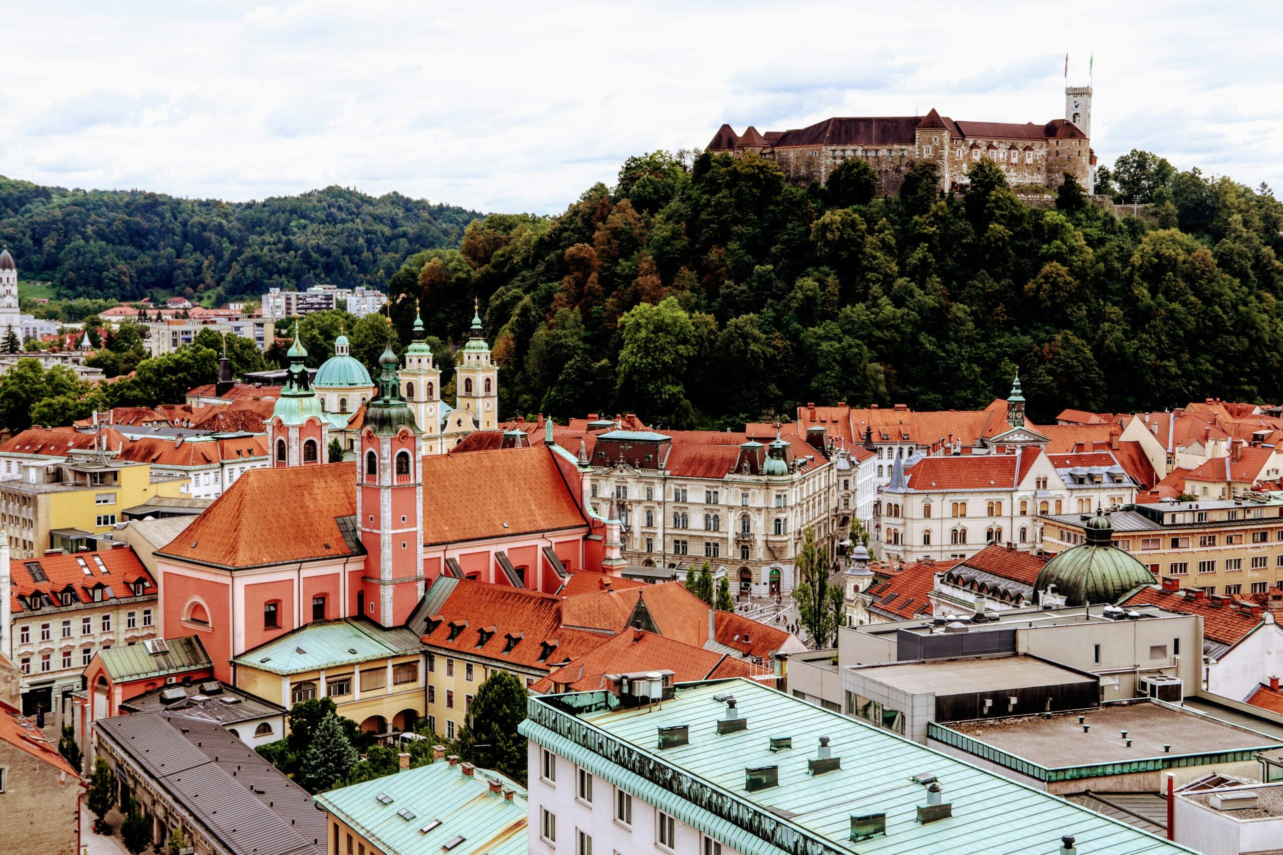 Pogled na Ljubljano z značilnim rdečim cerkvenim zvonikom v ospredju in Ljubljanskim gradom na zelenem hribu v ozadju