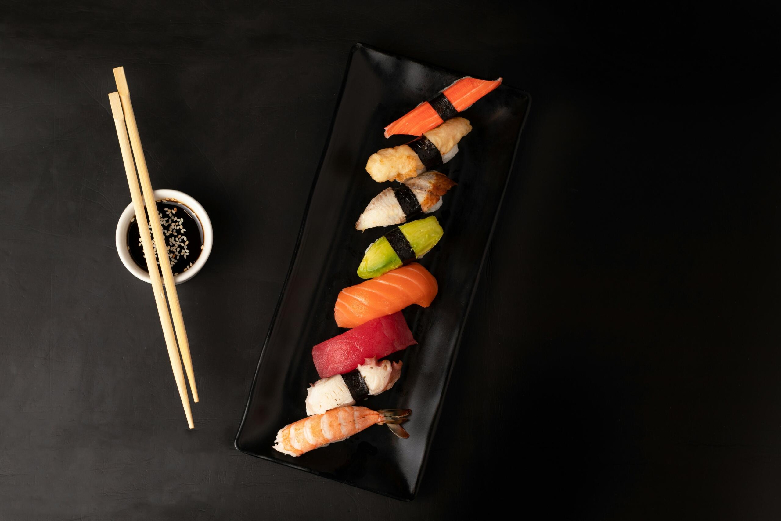 Eleganten niz nigiri sušija na dolgem črnem krožniku – tuna, losos, avokado, kozica in druge vrste, poleg pa palčke in sojina omaka