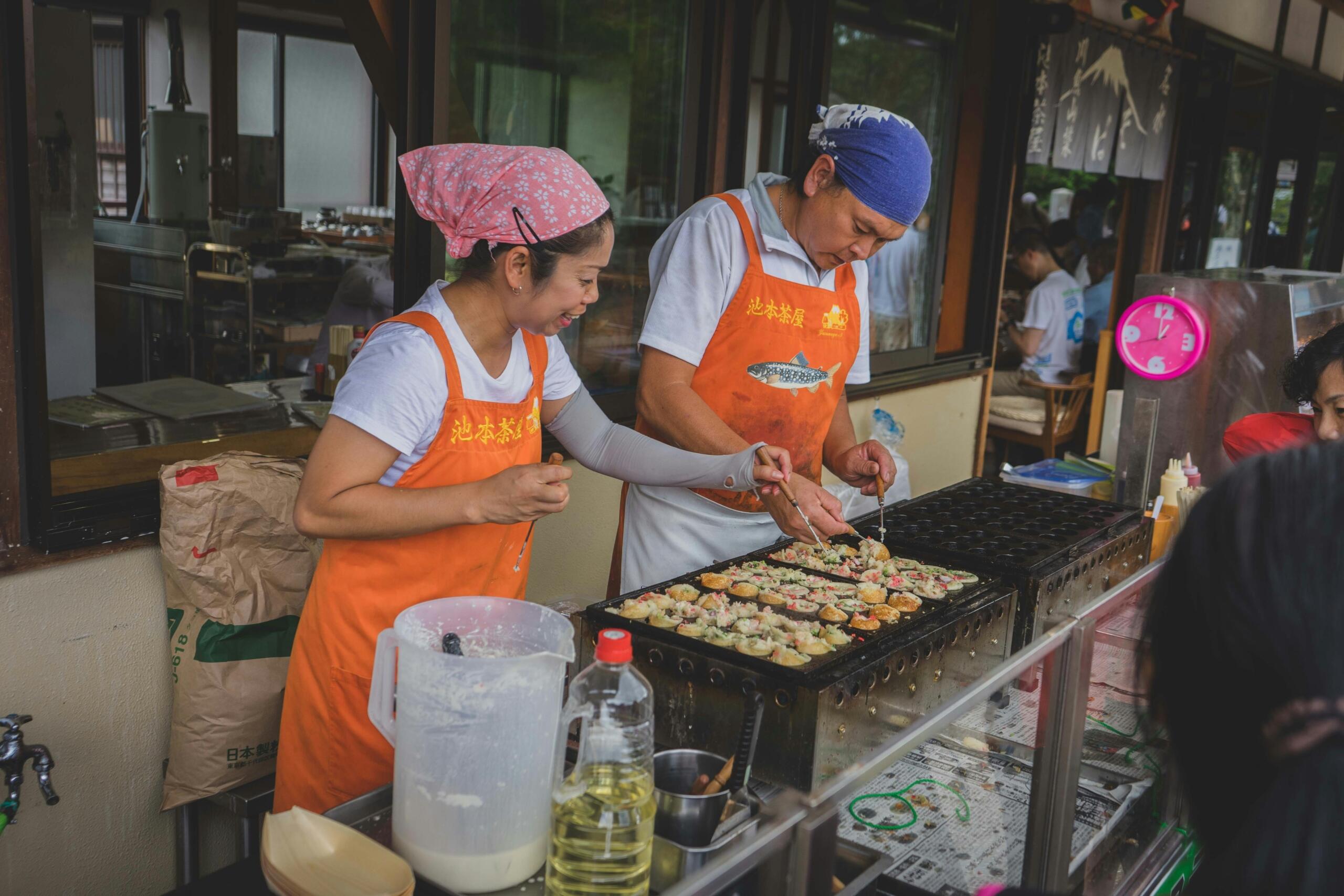 Moški in ženska v oranžnih predpasnikih skupaj pripravljata takoyaki na veliki okrogli plošči, ženska se smeji