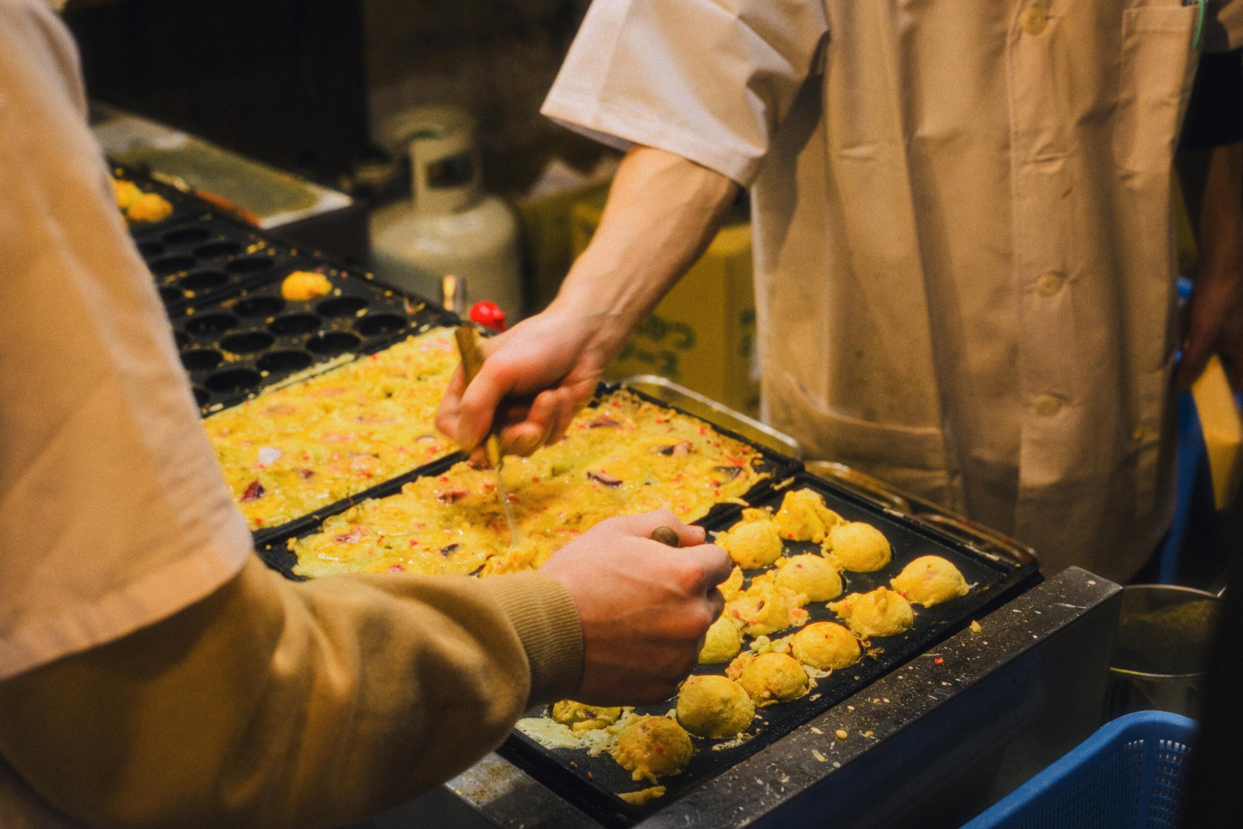 Roke kuharja z leseno palčko obračajo zlato-rumene takoyaki kroglice na črni plošči