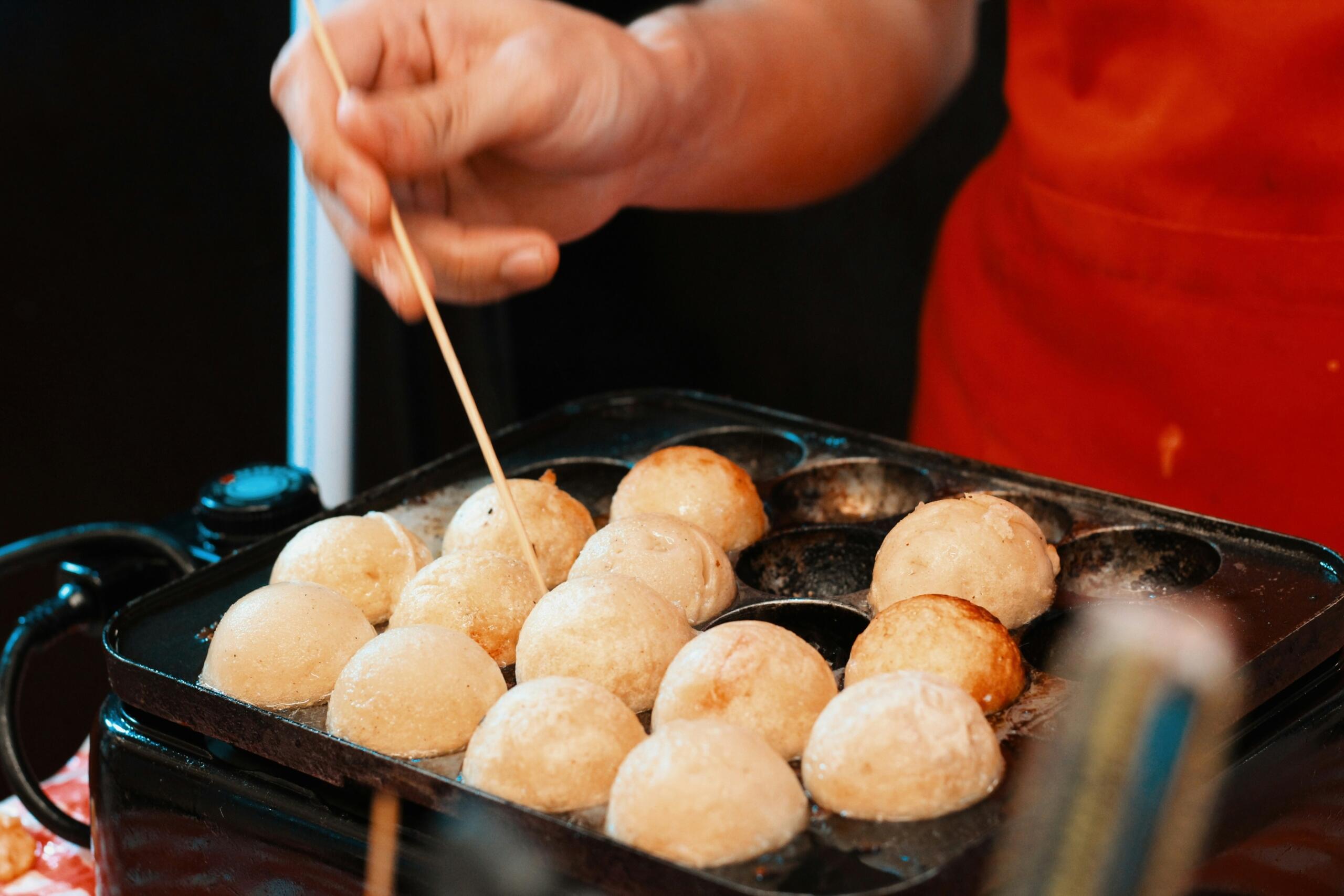 Blizu posnetek takoyaki kroglic na črni železni plošči, roka z leseno palčko jih previdno obrača