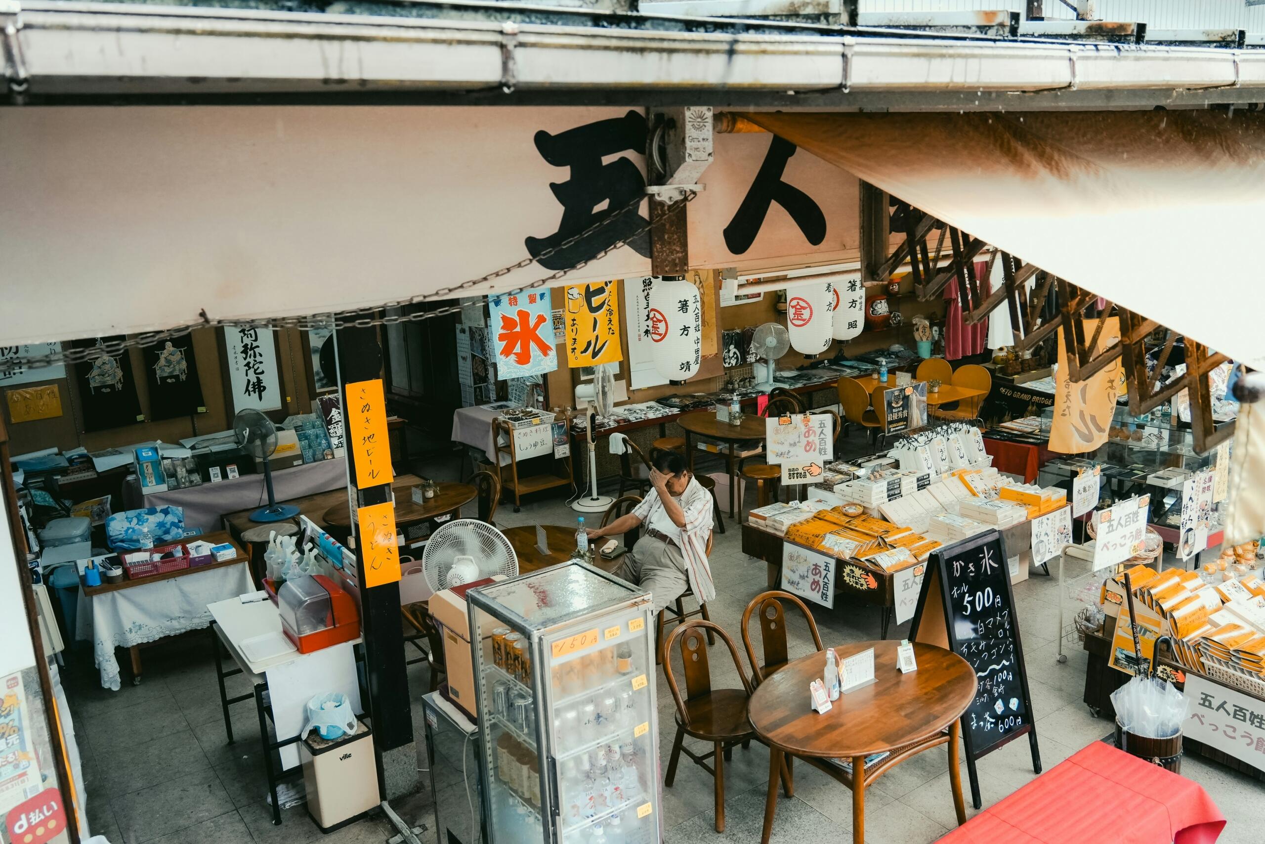Notranjost tradicionalne japonske trgovine in gostilne z lesenimi mizami, številnimi napisi, laternami in moškim, ki sedi za mizo