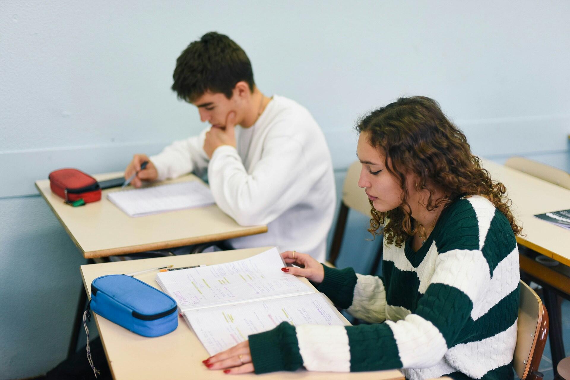 Študenti sedia v triede a riešia úlohy, dievča si číta poznámky, chlapec sa sústredene pozerá na test.
