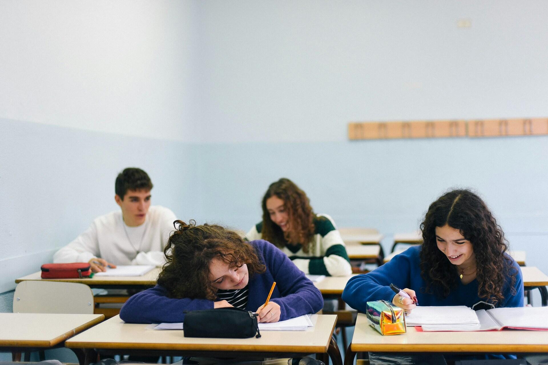 Skupina študentov píše test v triede, niektorí sa usmievajú, iní sú sústredení.