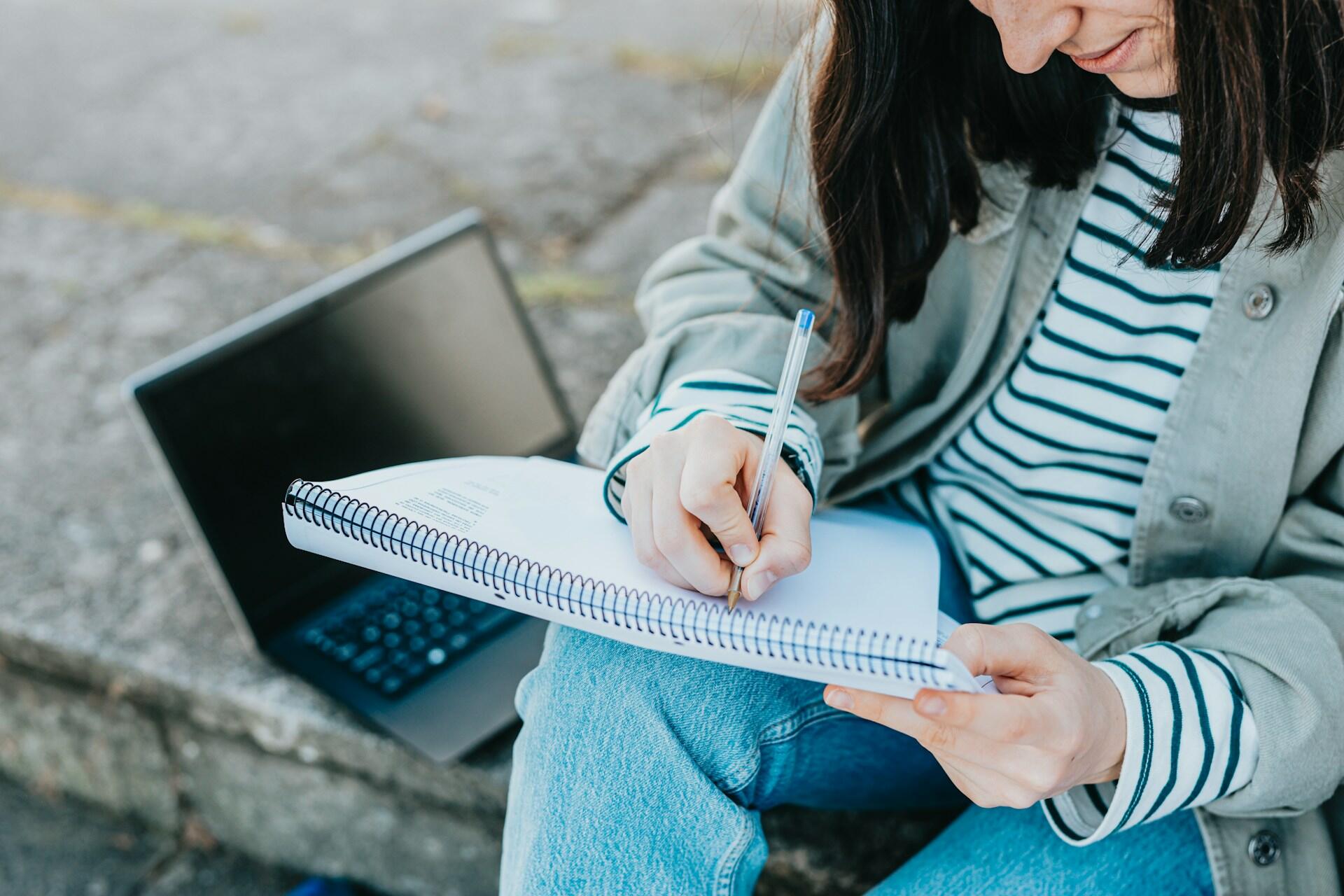 Študentka si zapisuje poznámky do zošita pri učení vonku s notebookom