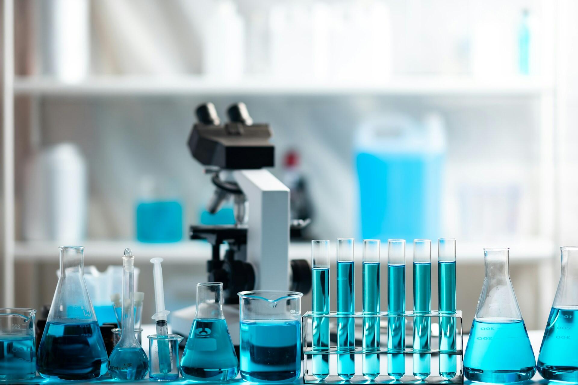 Skúmavky, banky a mikroskop na stole v chemickom laboratóriu s modrými roztokmi