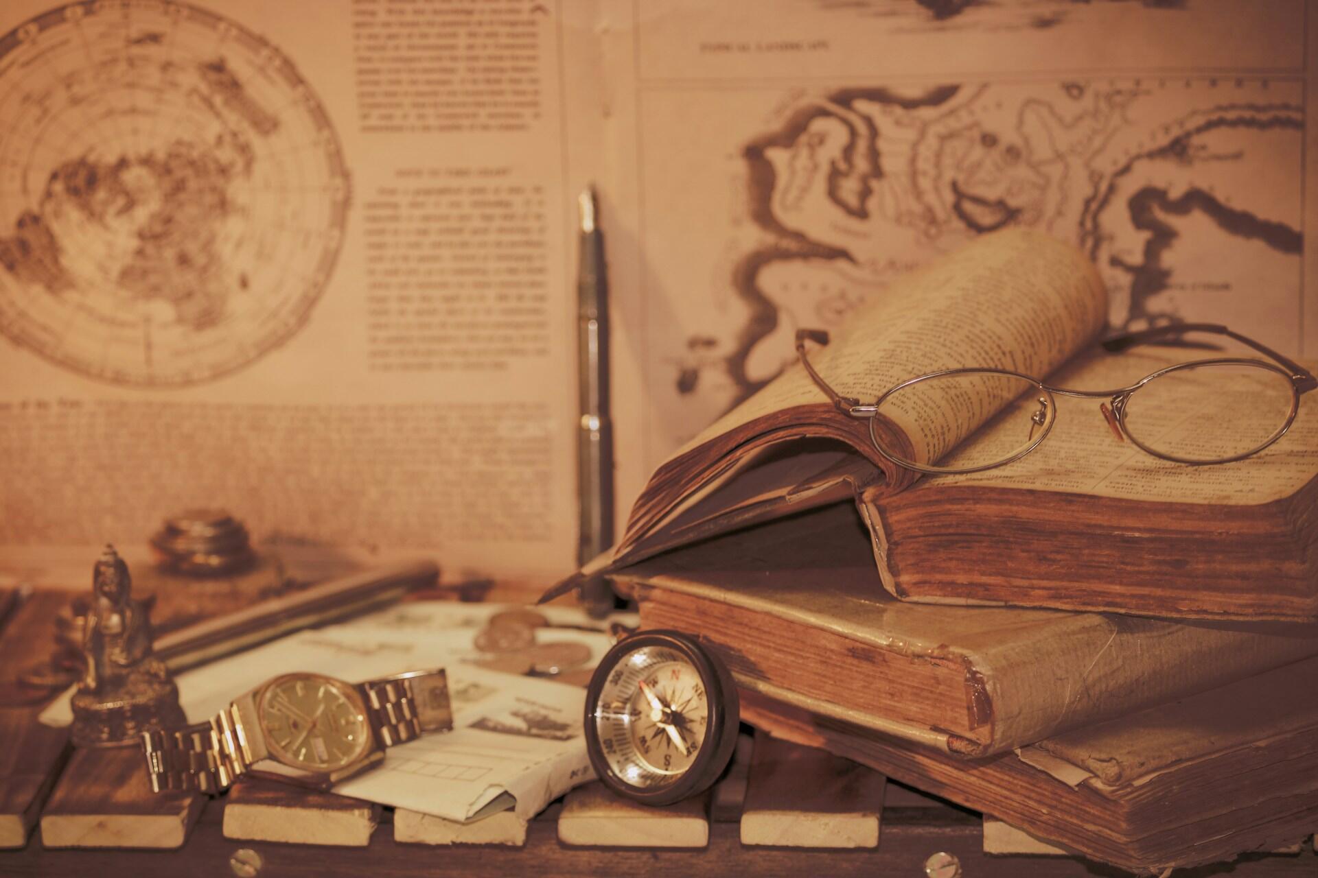 Staré knihy s okuliarmi, kompasom a historickými mapami na stole.