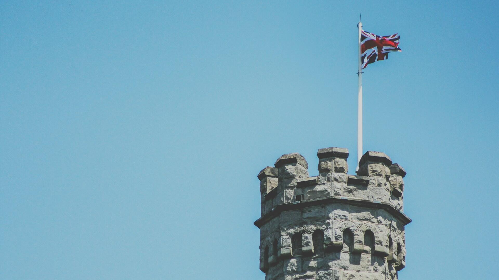 Britská vlajka vejúca na historickej kamennej veži pod modrou oblohou