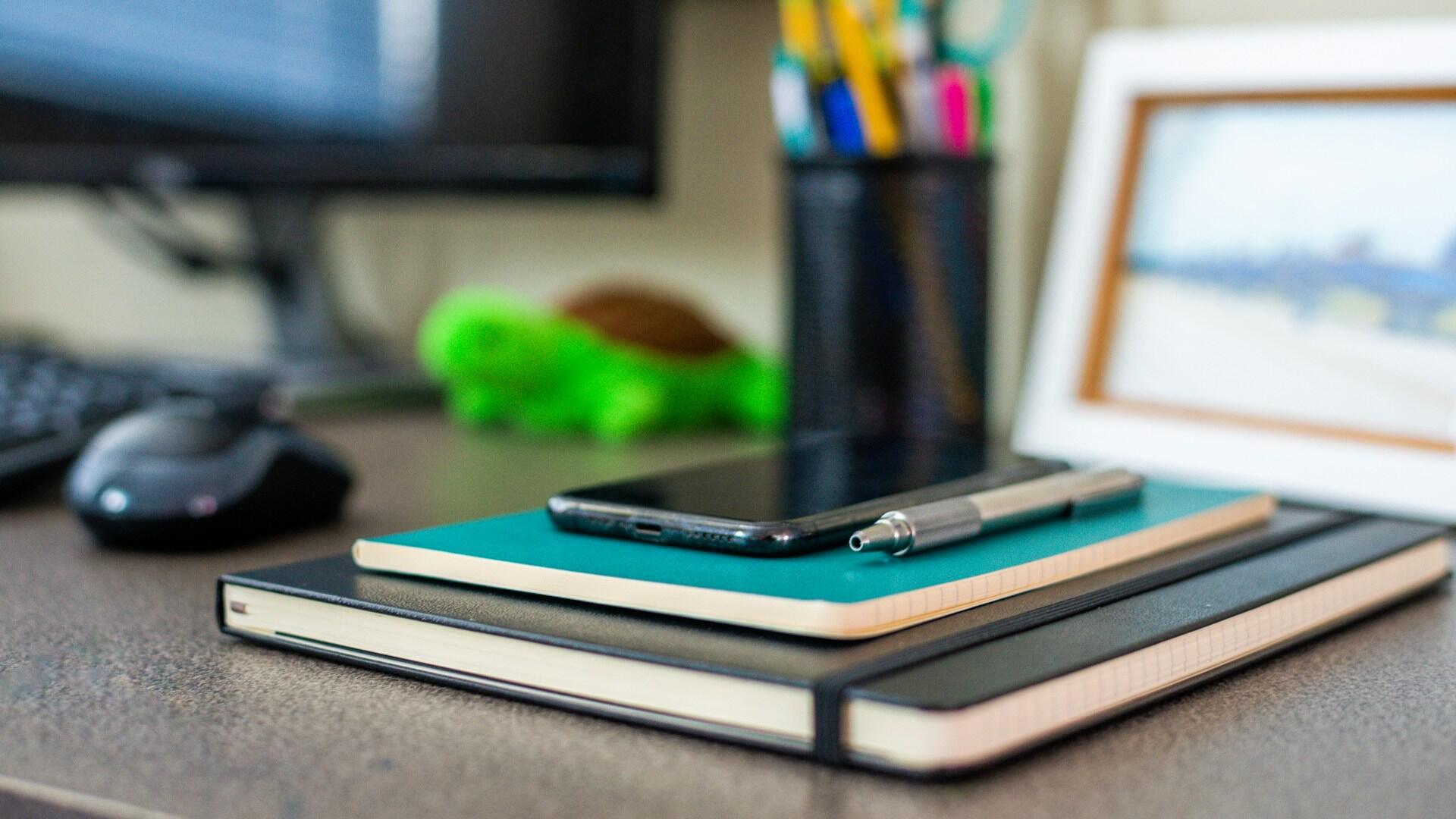 Zatvorené zápisníky so smartfónom a perom položené na pracovnom stole.