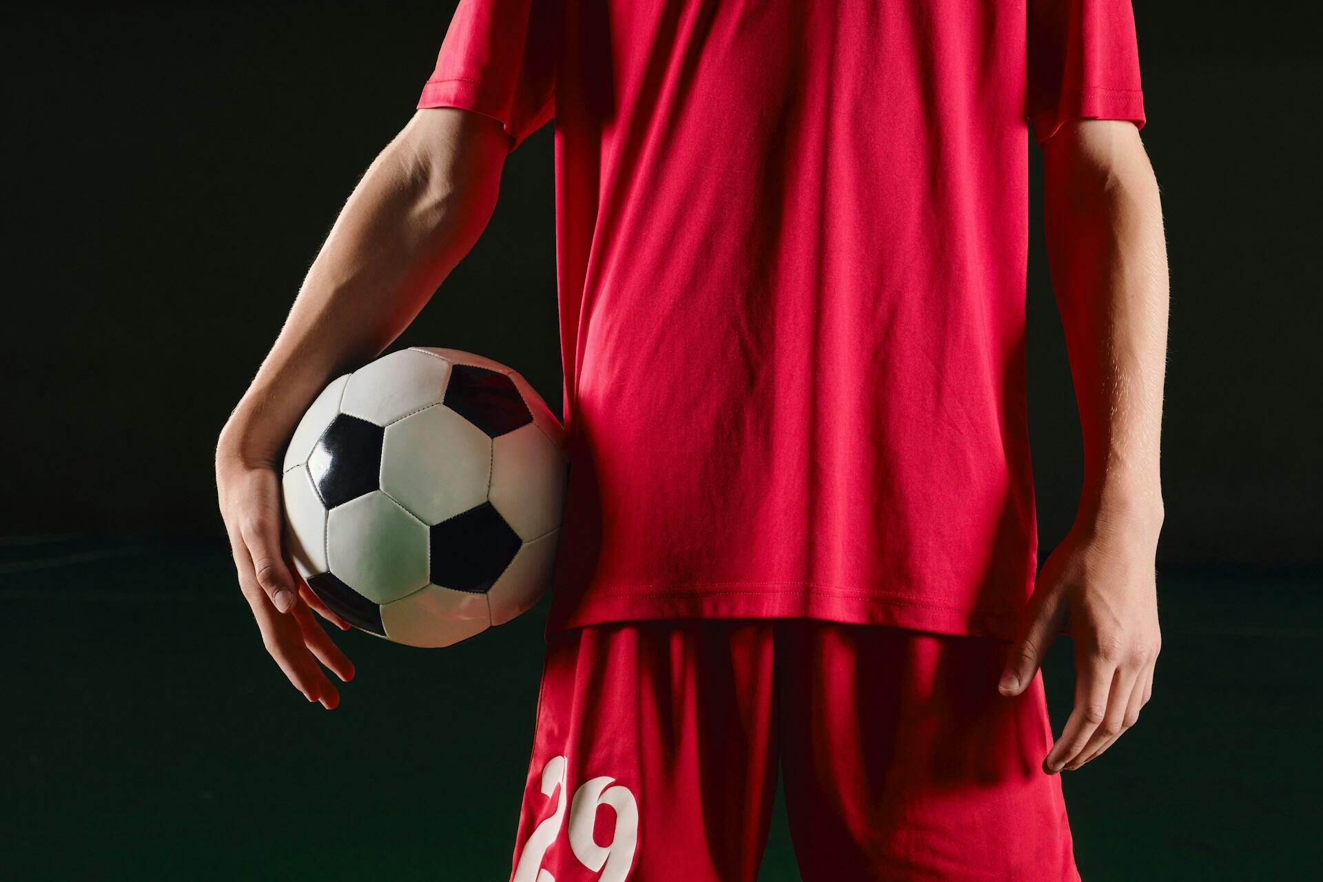 Futbalista v červenom drese drží futbalovú loptu pripravený na zápas