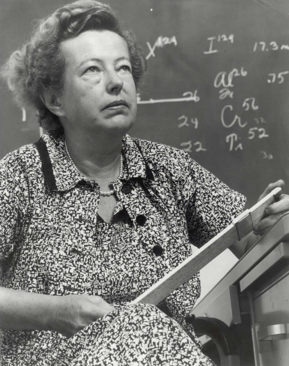 Maria Goeppert Mayer sedí pred tabuľou s fyzikálnymi výpočtami a drží meracie pravítko