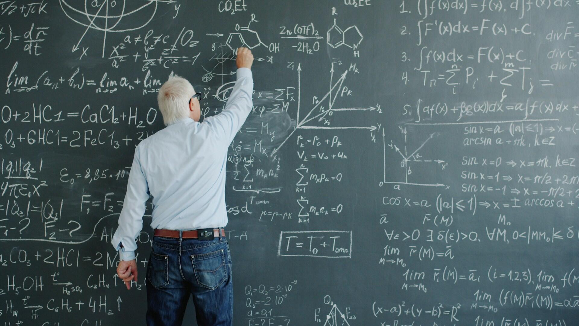 Profesor píše matematické a fyzikálne vzorce na tabuľu plnú rovníc